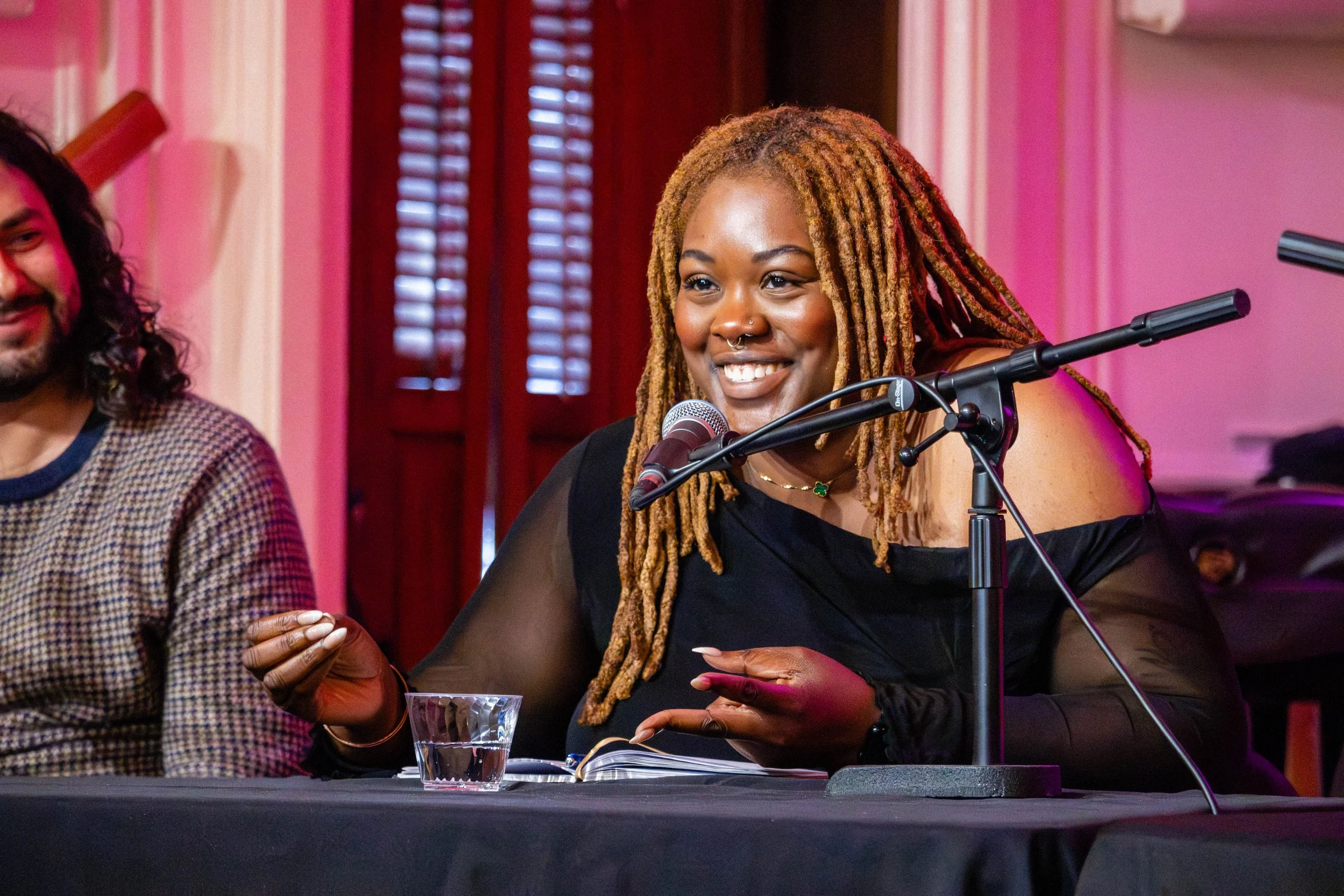 A woman with blonde dreadlocks smiling and sitting at a table with a glass of water, speaking into a microphone in a room with pink and brown walls and windows with wooden blinds.