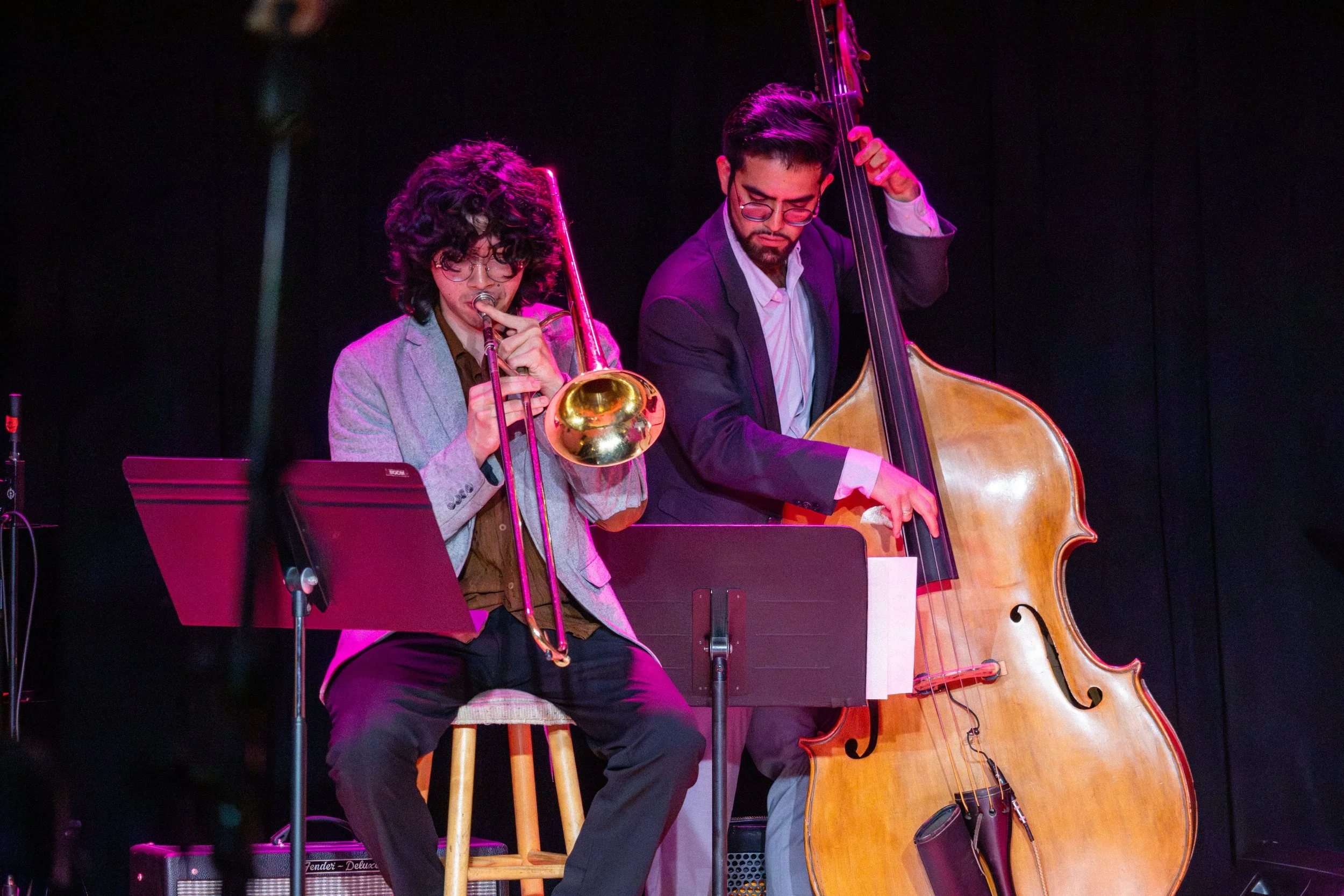 Two musicians performing on stage, a trumpet player on the left and a double bassist on the right. Both are dressed in suits and are focused on their instruments, with stage lighting highlighting them against a dark background.