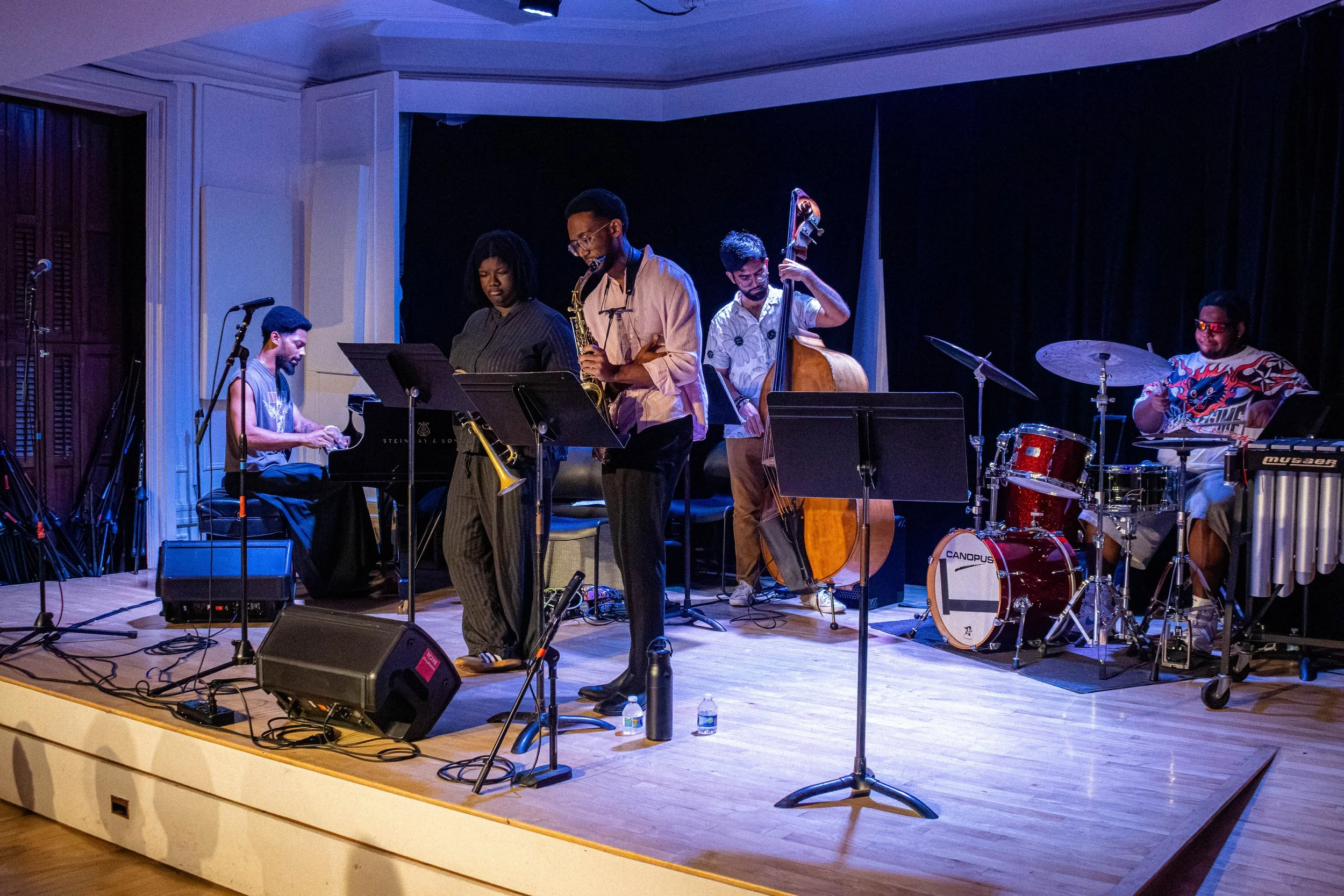 A jazz band performing on a stage with five musicians playing various instruments, including piano, saxophone, double bass, drums, and vibraphone, under stage lighting.