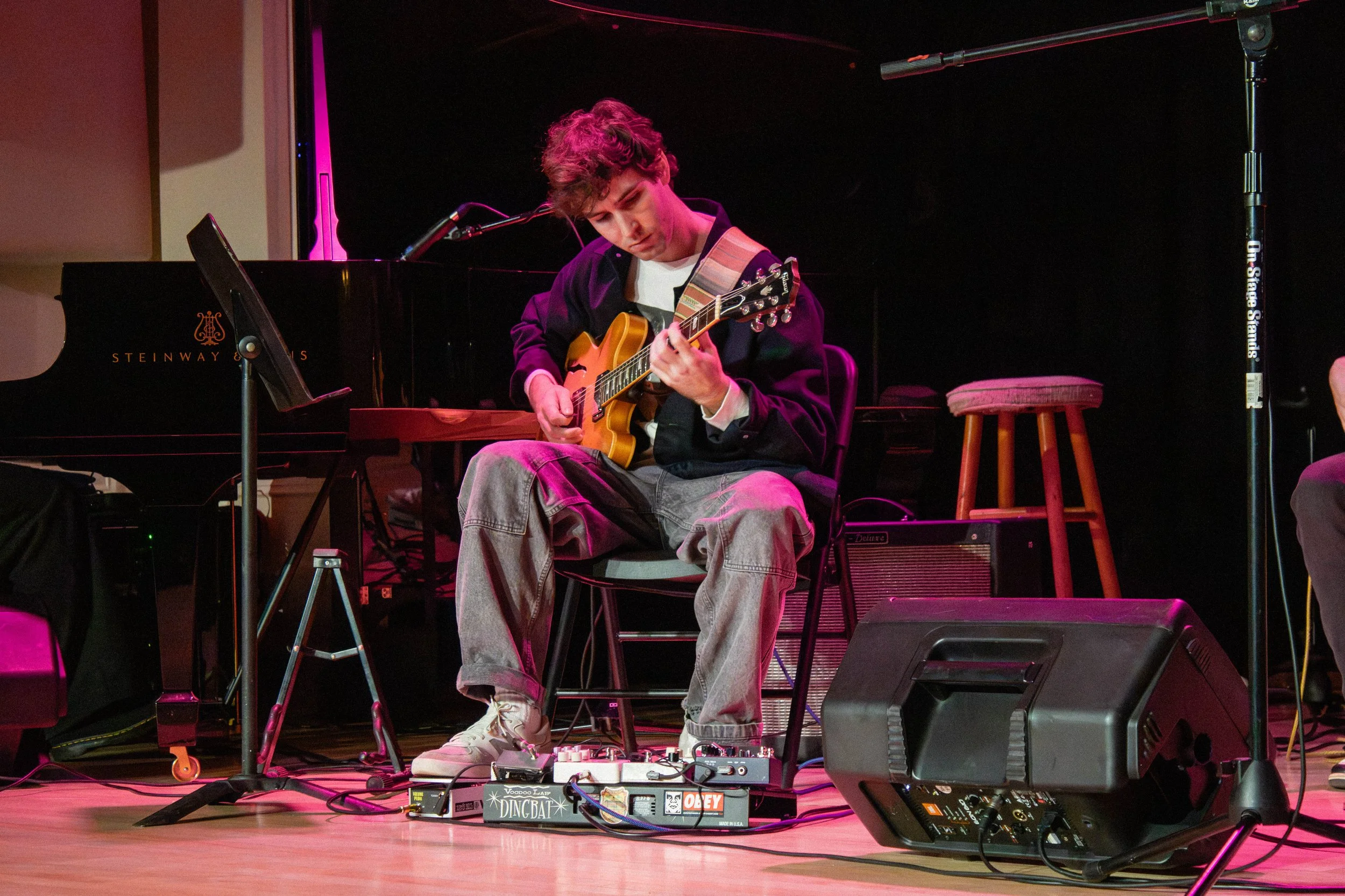 A young man plays an electric guitar on a stage, sitting on a chair. There is a black Steinway grand piano behind him, a stool to his right, and various musical equipment and pedals on the floor in front of him. The setting is a dimly lit performance