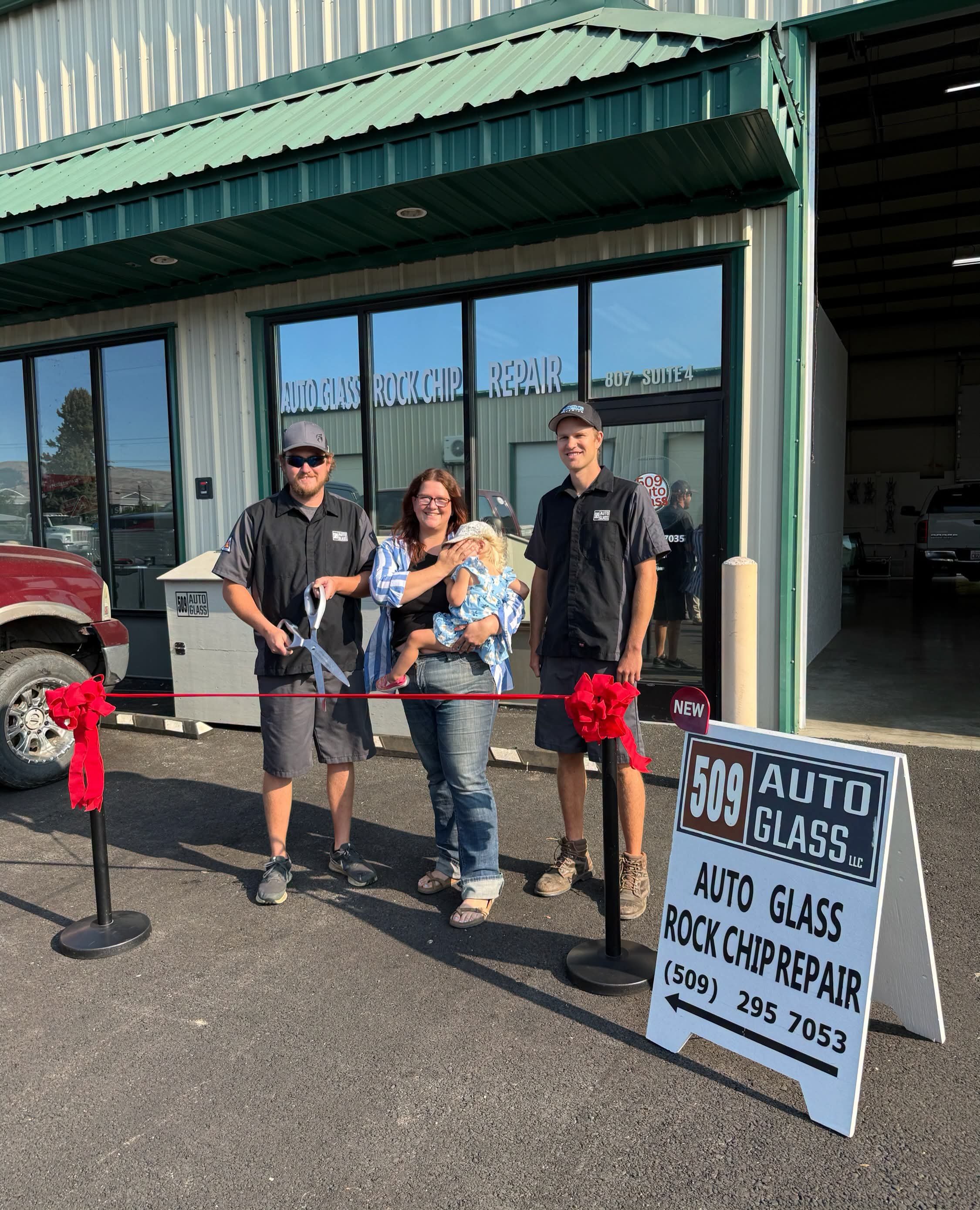 A group of four people, including a woman holding a young girl, standing outside a building with a sign for auto glass and rock chip repair. There is a red ribbon in front of them, indicating a ribbon-cutting ceremony. The building has large glass windows with the business name and contact information visible. Two men are dressed in black work uniforms, and the background includes other machinery and vehicles.