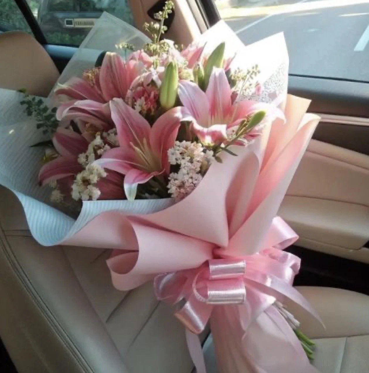 Een bouquet bloemen met roze lelies, witte bloemen en groene bladeren, ingepakt in roze en wit papier met een grote roze strik, neergelegd op een autostoel.