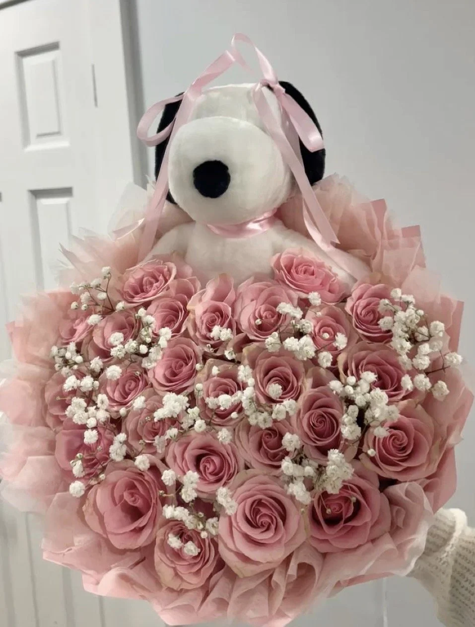 Een snoopy knuffel met een bloemenboeket van roze rozen en witte kleine bloemen, versierd met een roze lint, gehouden door een persoon met een witte gebreide handschoen.