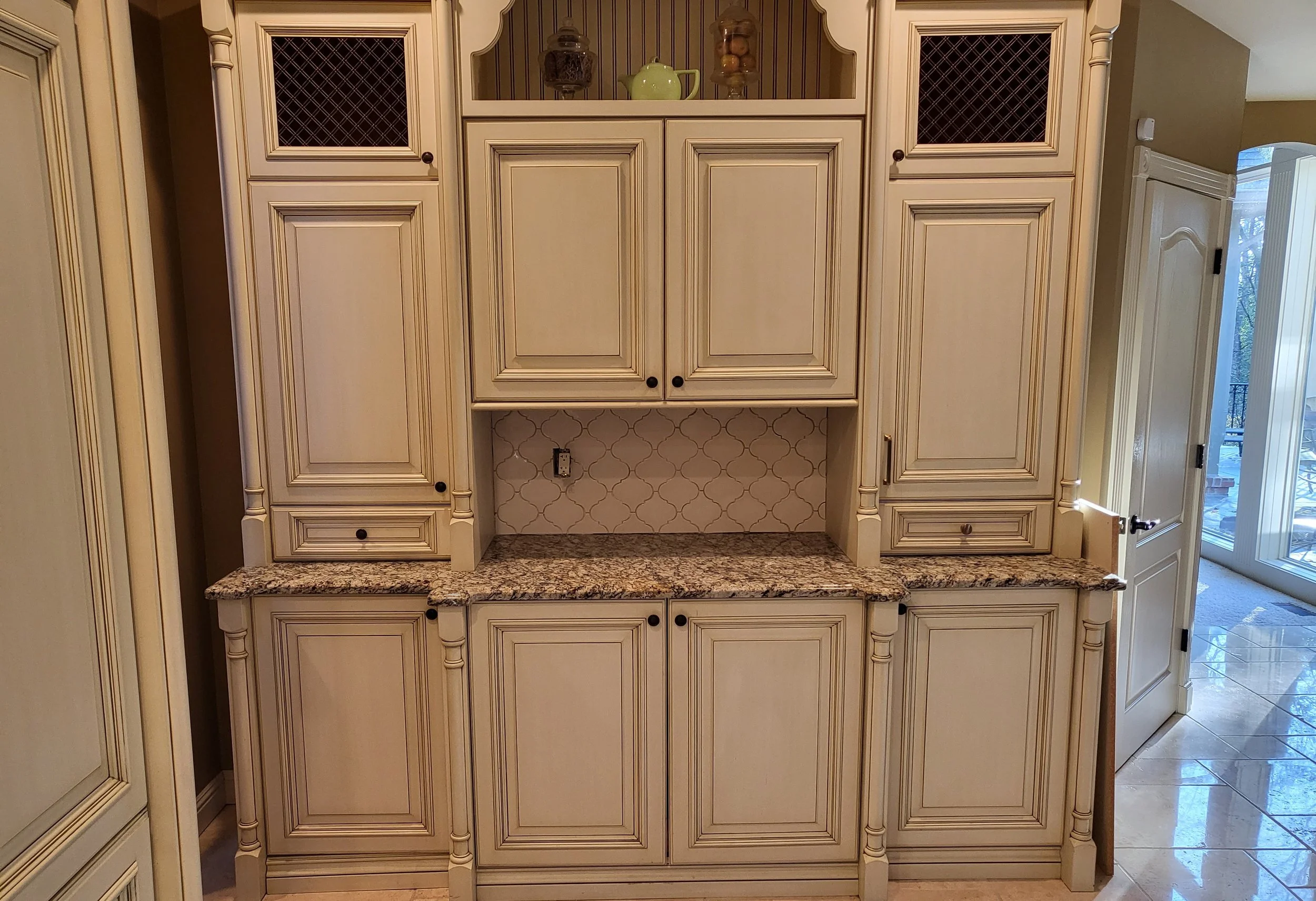 Cream-colored kitchen cabinets with detailed molding, black knobs, granite countertop, and a tiled backsplash with a quatrefoil pattern.