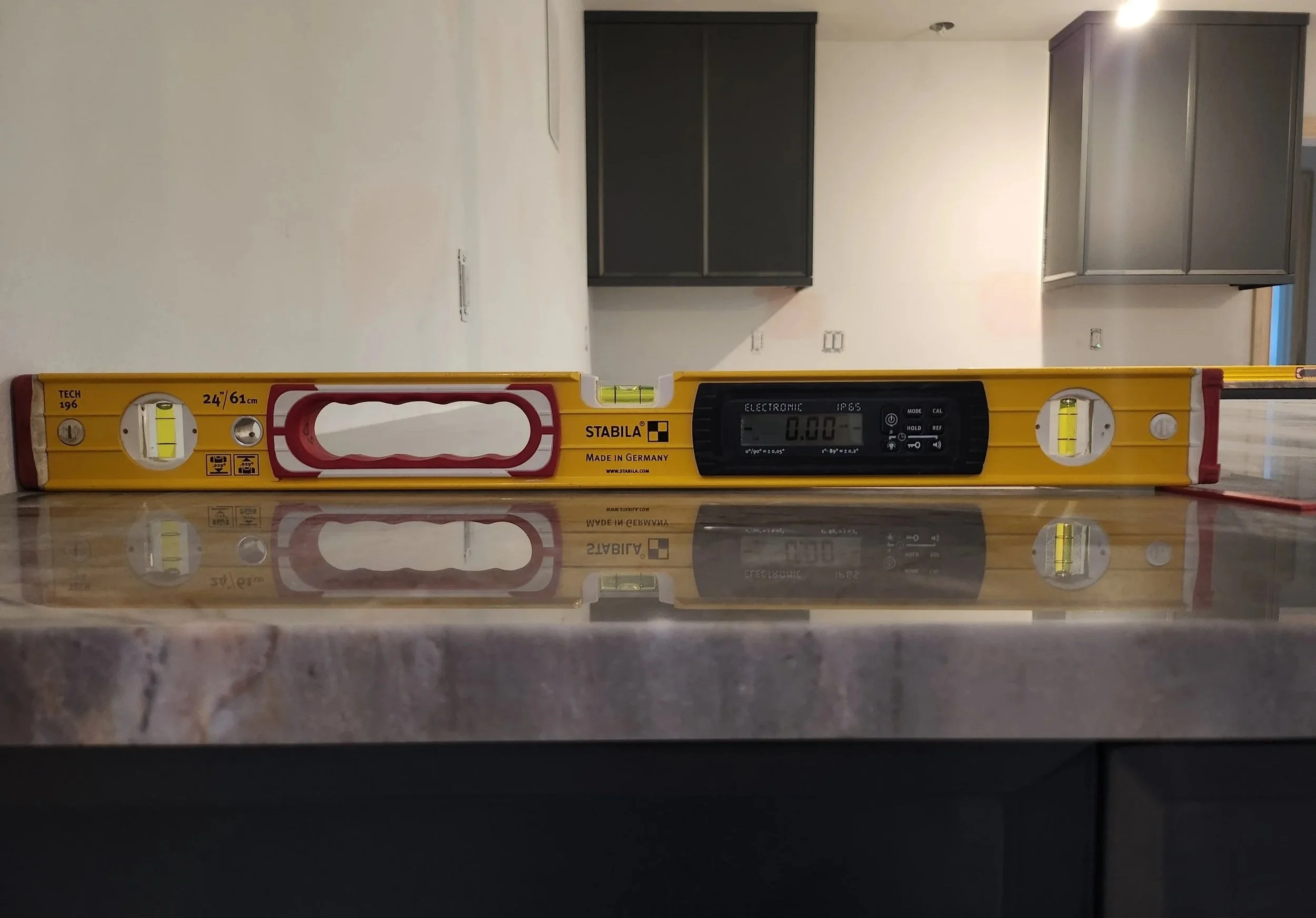 A yellow spirit level with black digital readout placed on a dark surface with a reflection, against a background of an unfinished room with white walls and dark cabinets.
