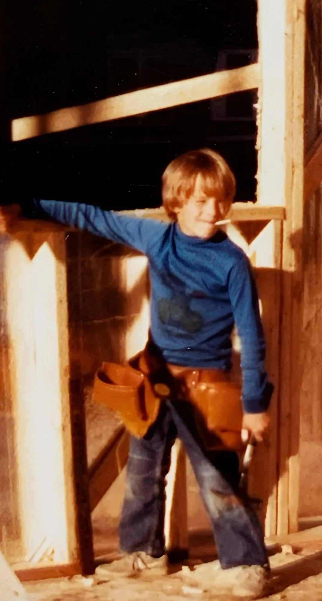 A young boy standing in a room under construction, wearing a blue long sleeve shirt, jeans, and a brown tool belt.