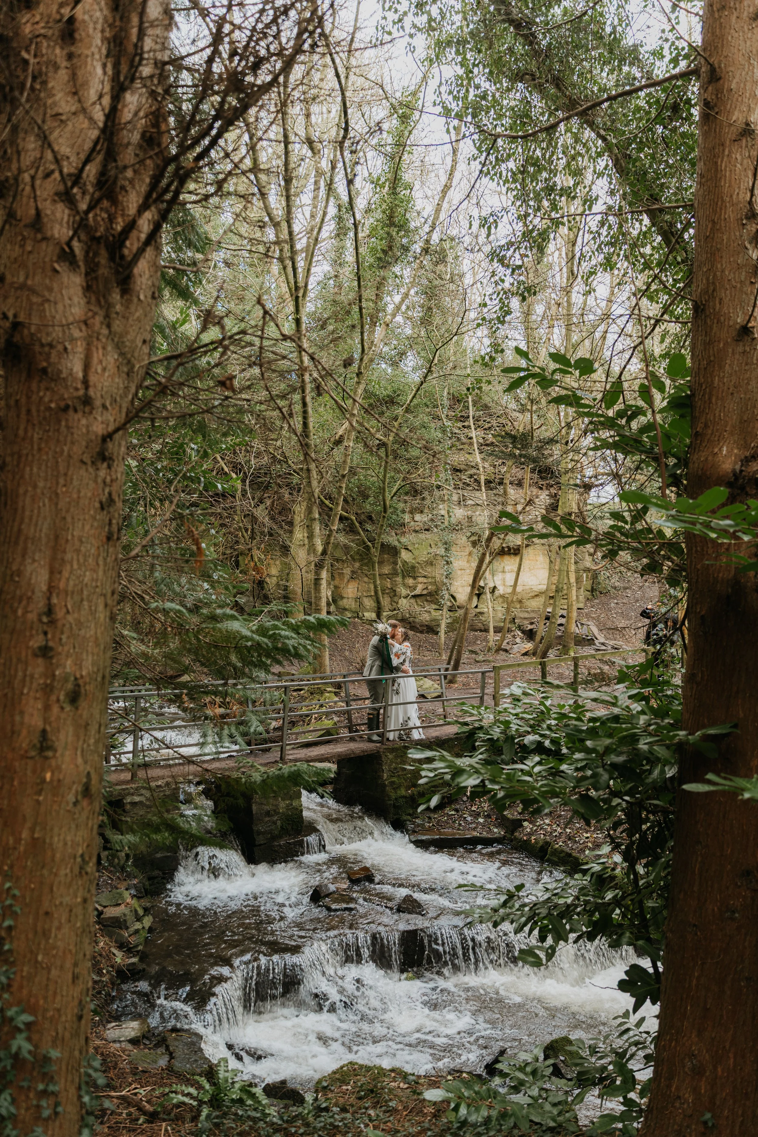 A couple in wedding attire standing on a small bridge over a stream in a wooded area, surrounded by trees and foliage. The Garden Rooms at Tennants, Leyburn, North Yorkshire