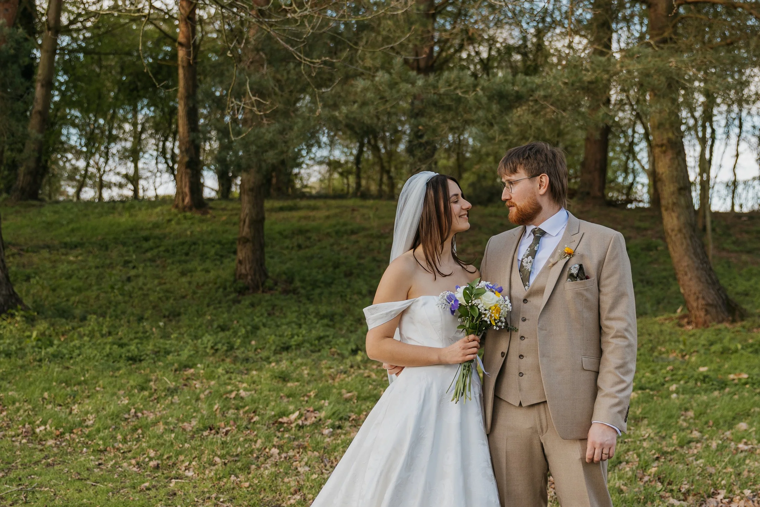A bride and groom standing close together outdoors in a wooded area, smiling at each other. The bride wears a white wedding dress and a veil, holding a bouquet of flowers. The groom wears a beige suit and glasses.