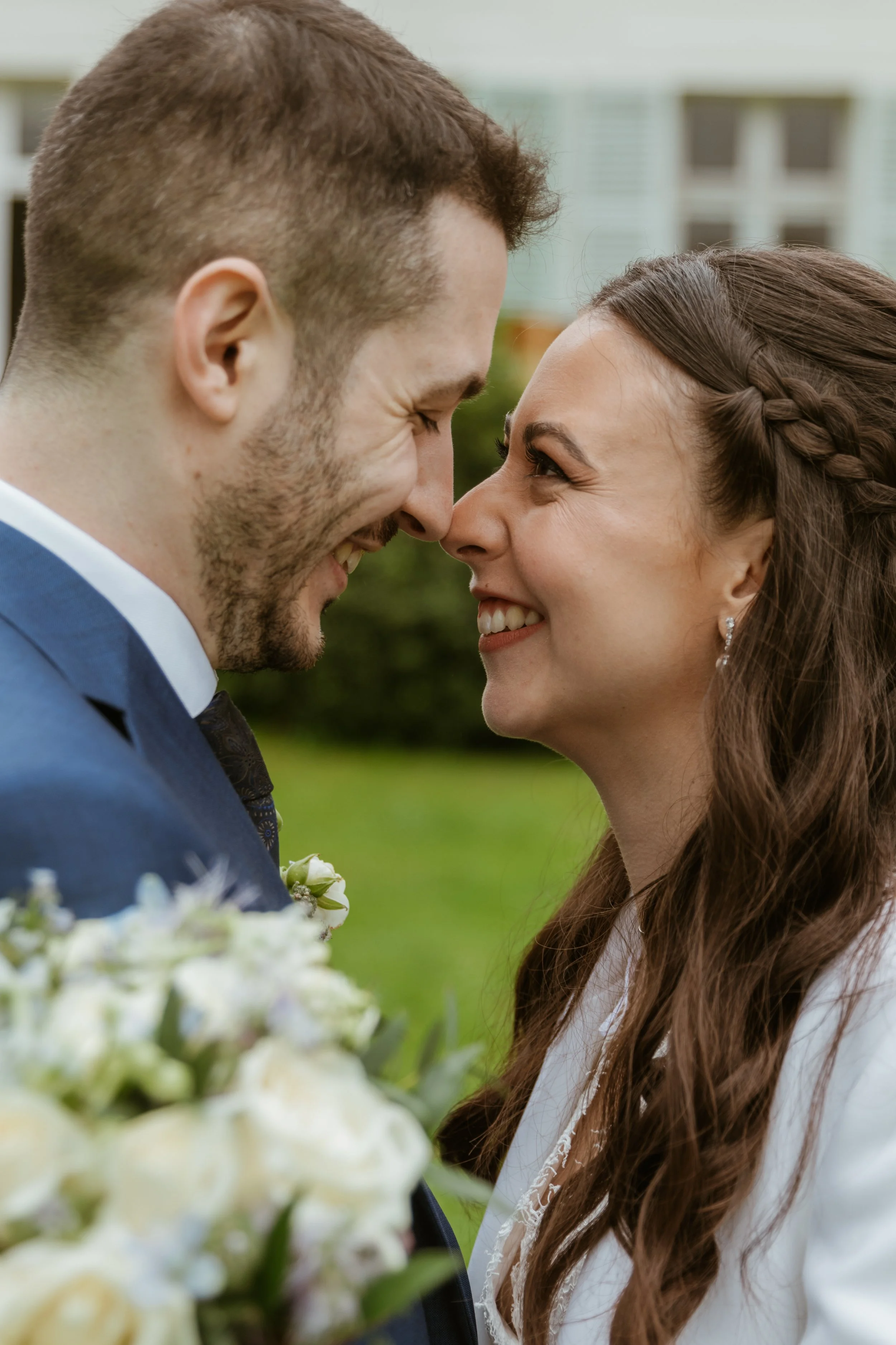 A couple faces each other closely outdoors, smiling and about to kiss, with blurred greenery and house in background.