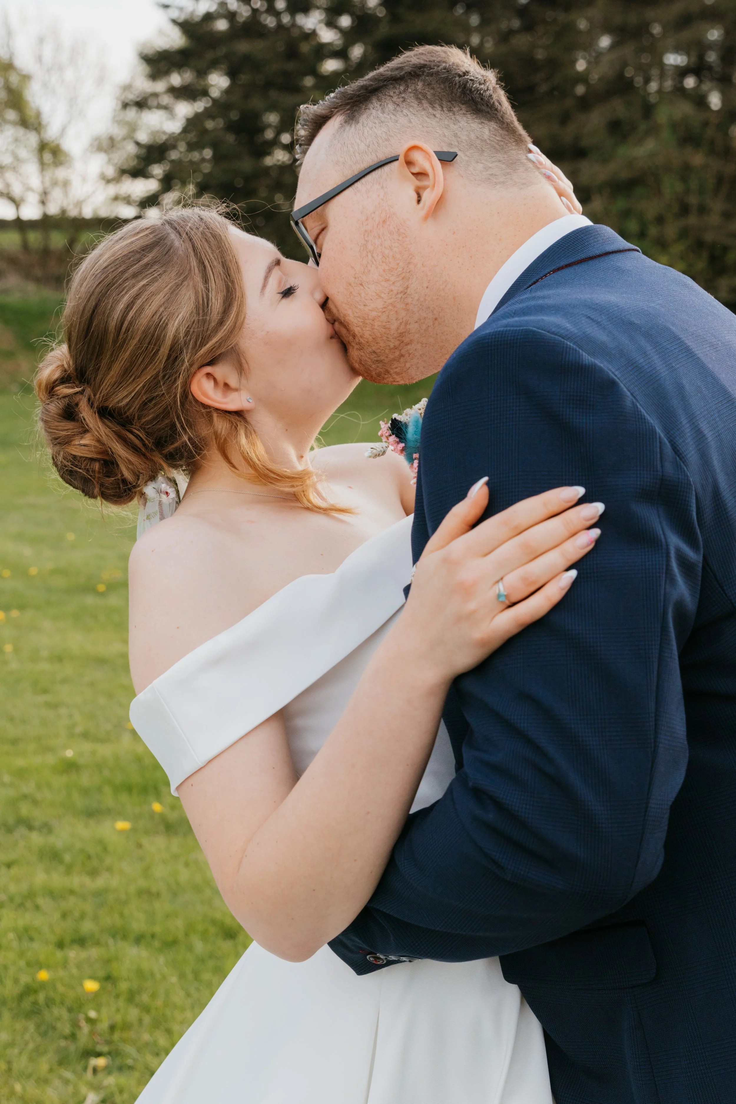 A man and a woman are kissing outdoors on a grassy area with trees in the background. The woman has light brown hair styled in a braid and is wearing a white dress. The man wears glasses, a blue suit, and a white shirt.