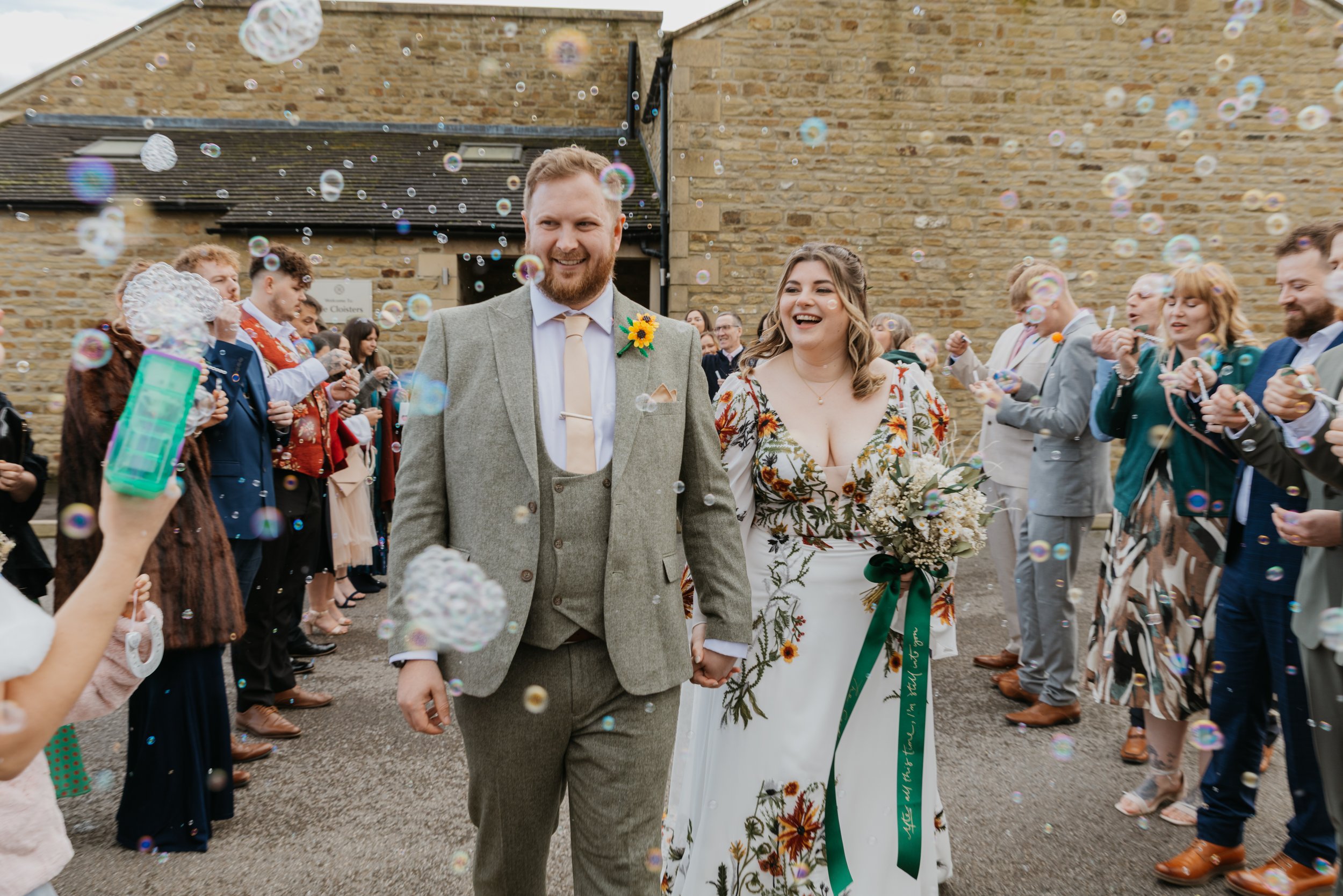A happy bride and groom walking hand in hand outside a building with a crowd celebrating and blowing bubbles around them. The Garden Rooms at Tennants, Leyburn, North Yorkshire
