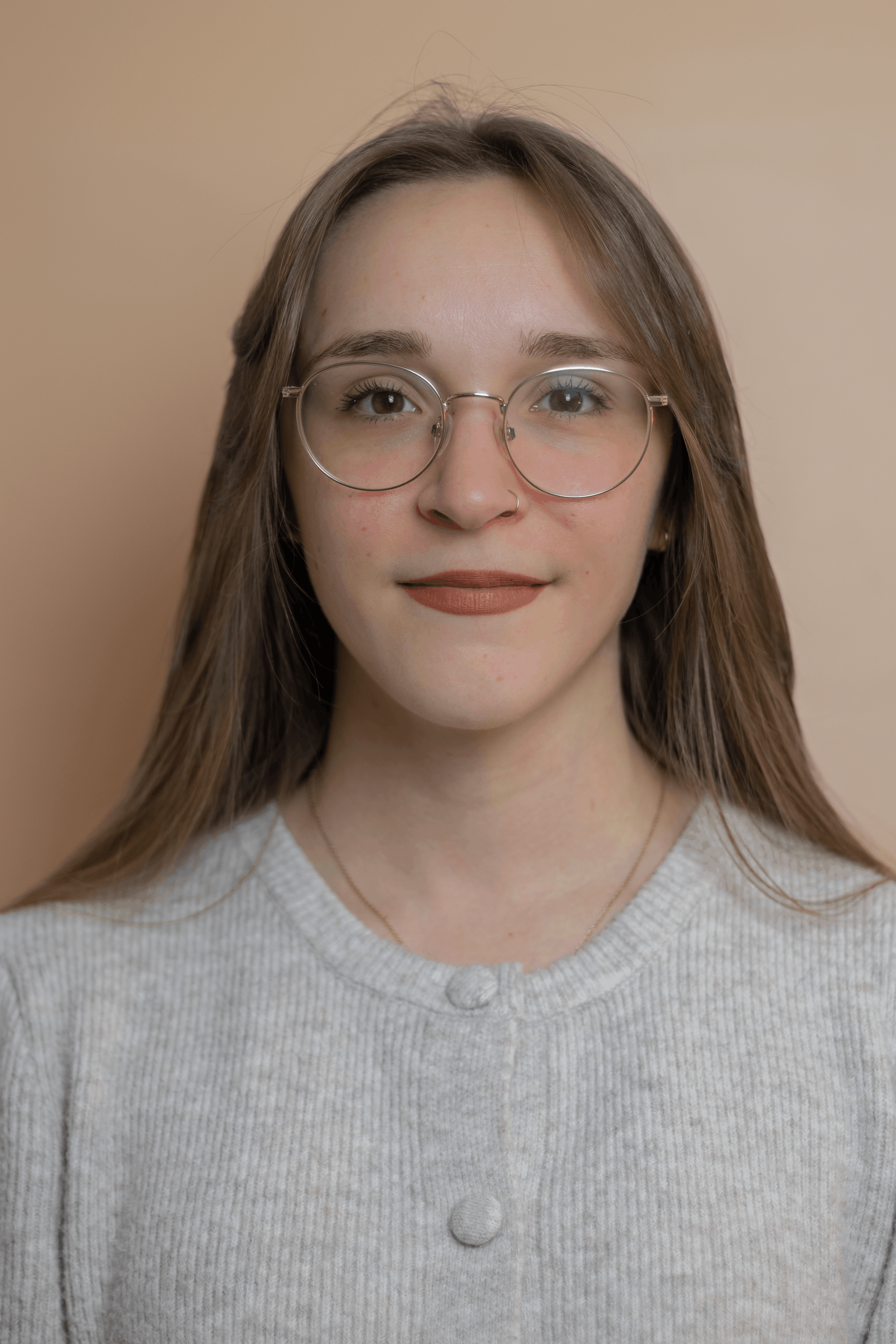Close-up of a woman with glasses, light brown hair, wearing a light gray buttoned cardigan and a gold necklace, against a beige background.