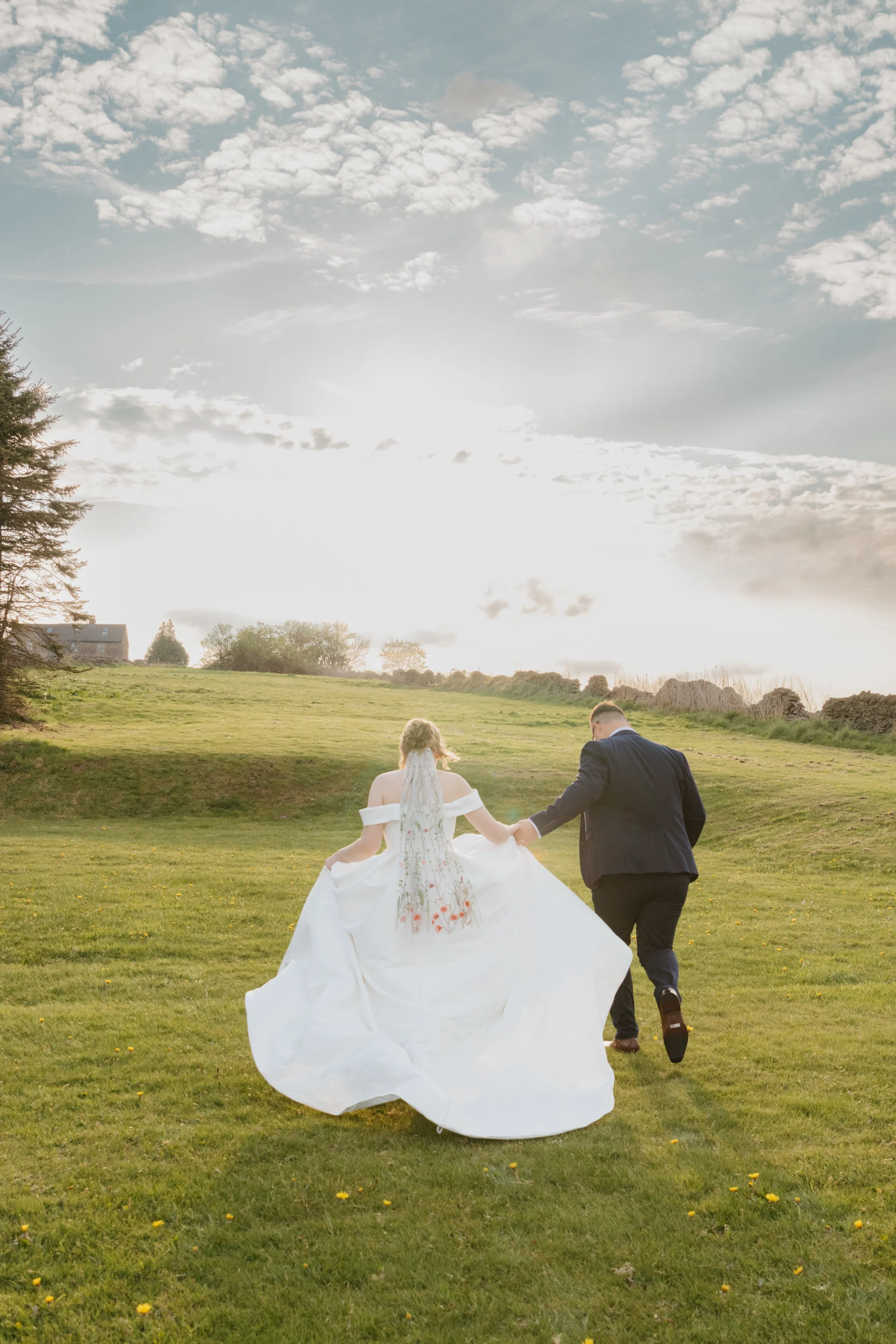 A couple dressed in wedding attire walking hand in hand on a lush green field with small yellow flowers, with the bride in a white gown with floral embroidery, and the groom in a dark suit, under a partly cloudy sky at sunset. Pudding Pie Hill