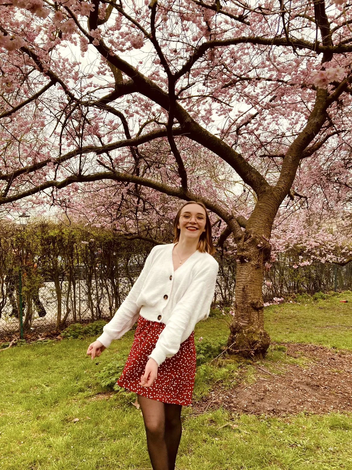 A young woman with glasses and shoulder-length hair smiling and standing outdoors in front of a flowering cherry blossom tree. She is wearing a white cardigan, a red skirt with white polka dots, and black tights.