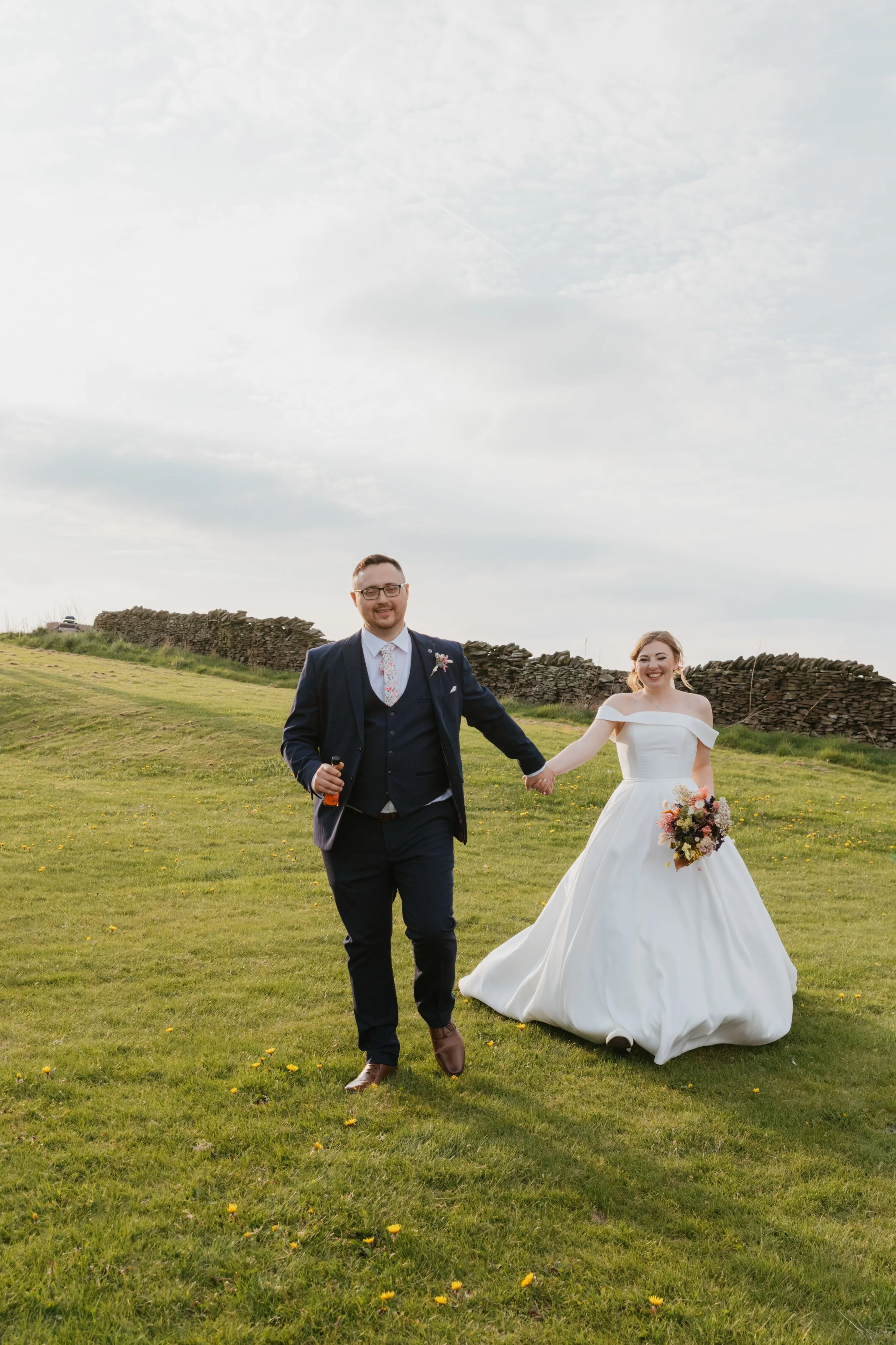 A bride and groom holding hands and walking outdoors on a grassy field with a stone wall behind them, smiling. The groom is wearing glasses, a navy blue suit, and a floral tie, while the bride is in a white wedding dress holding a bouquet of flowers.