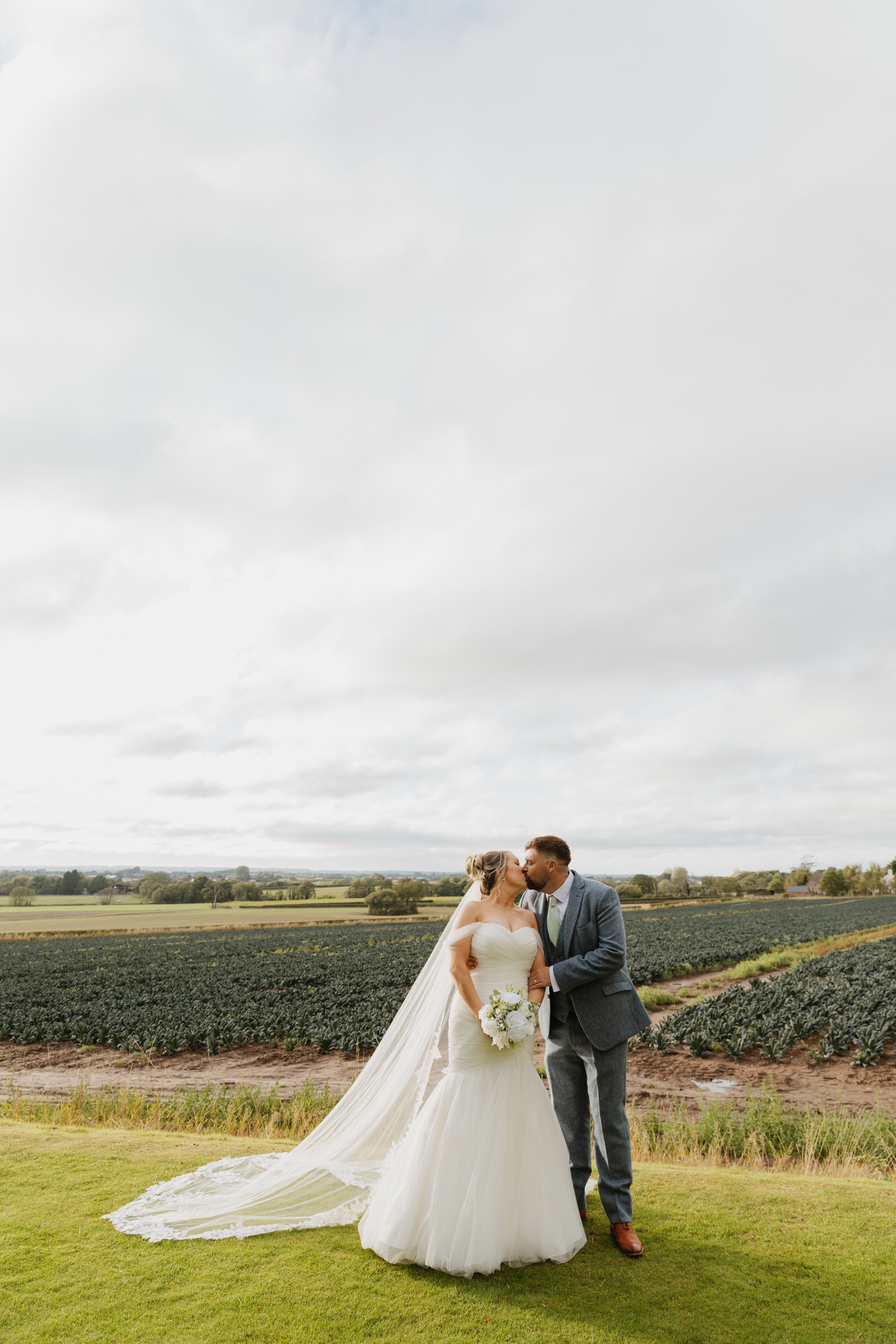 A bride and groom sharing a kiss outdoors in a rural field during their wedding, with green fields and trees in the background under a cloudy sky.