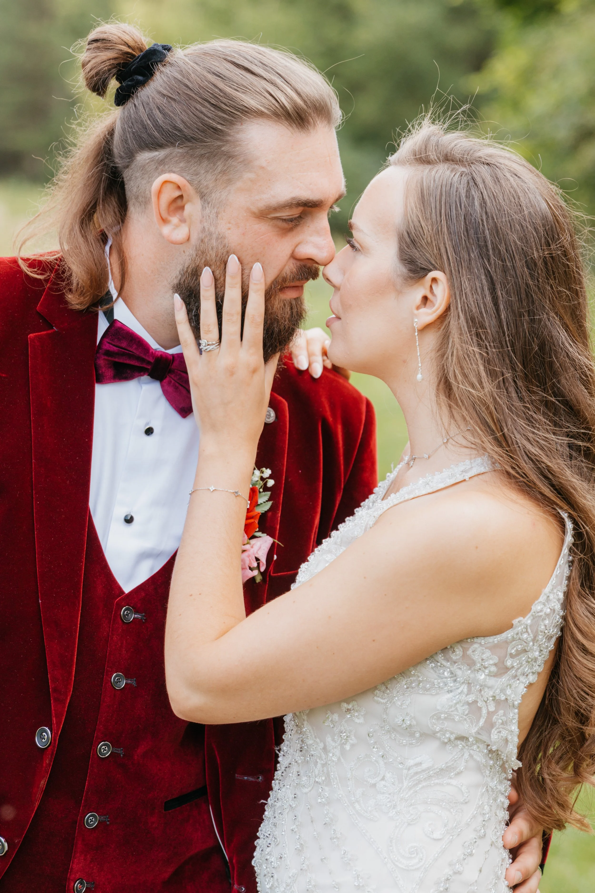 A bride and groom are close together, touching noses, outdoors with greenery in the background. The groom is wearing a red velvet suit with a bow tie and has long hair tied back with a black scrunchie, and a beard. The bride has long light brown hair, is wearing earrings, and a white lace wedding dress. The groom's left hand, showing a wedding ring, touches the bride's face.