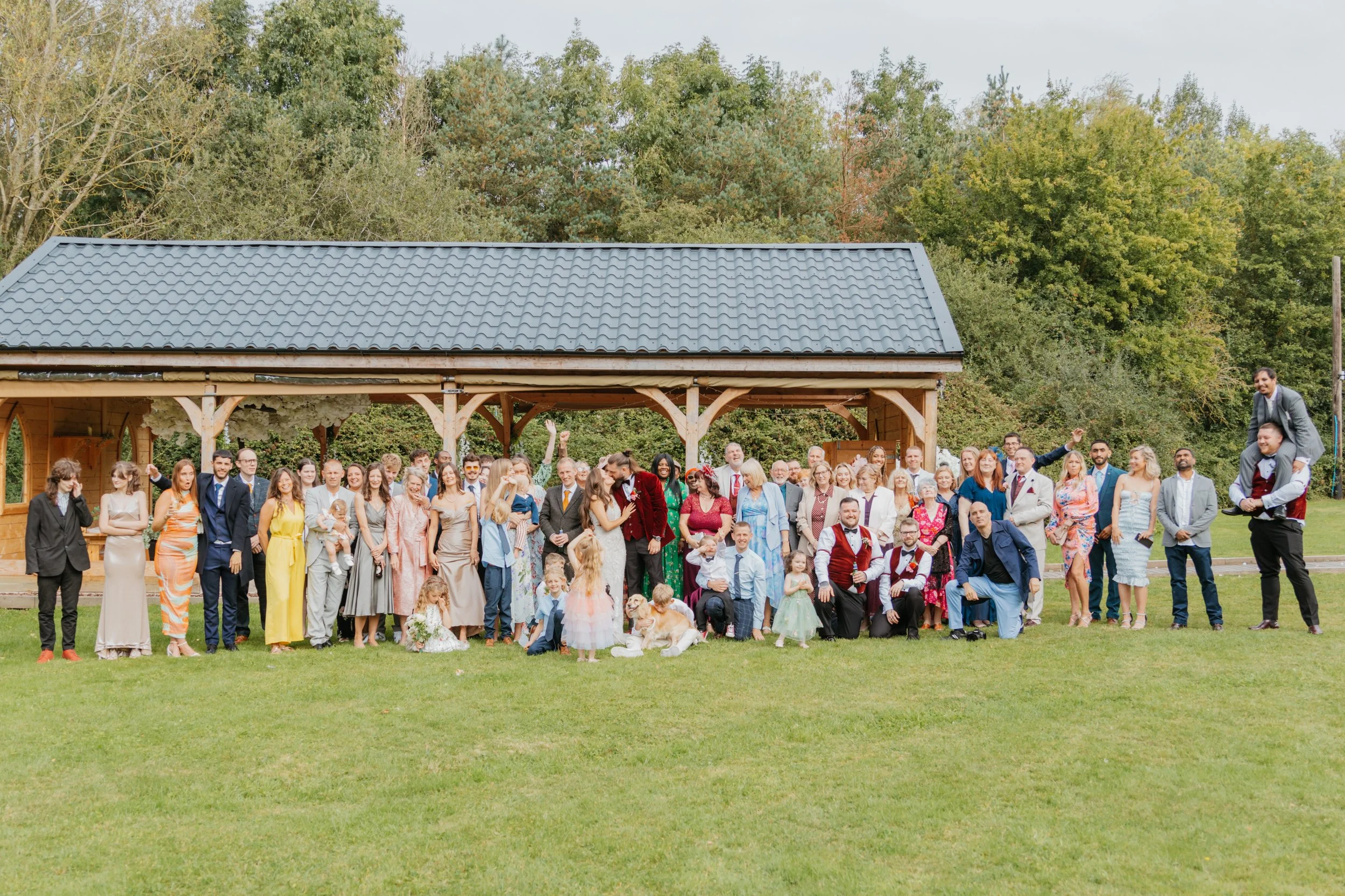 A large group of people gathered outdoors on a grassy area in front of a wooden pavilion with a dark tiled roof, surrounded by trees. The group includes adults and children, some dressed formally, some casually, and some in wedding attire. There is a man giving a piggyback ride, and people are smiling, waving, and posing for the camera.