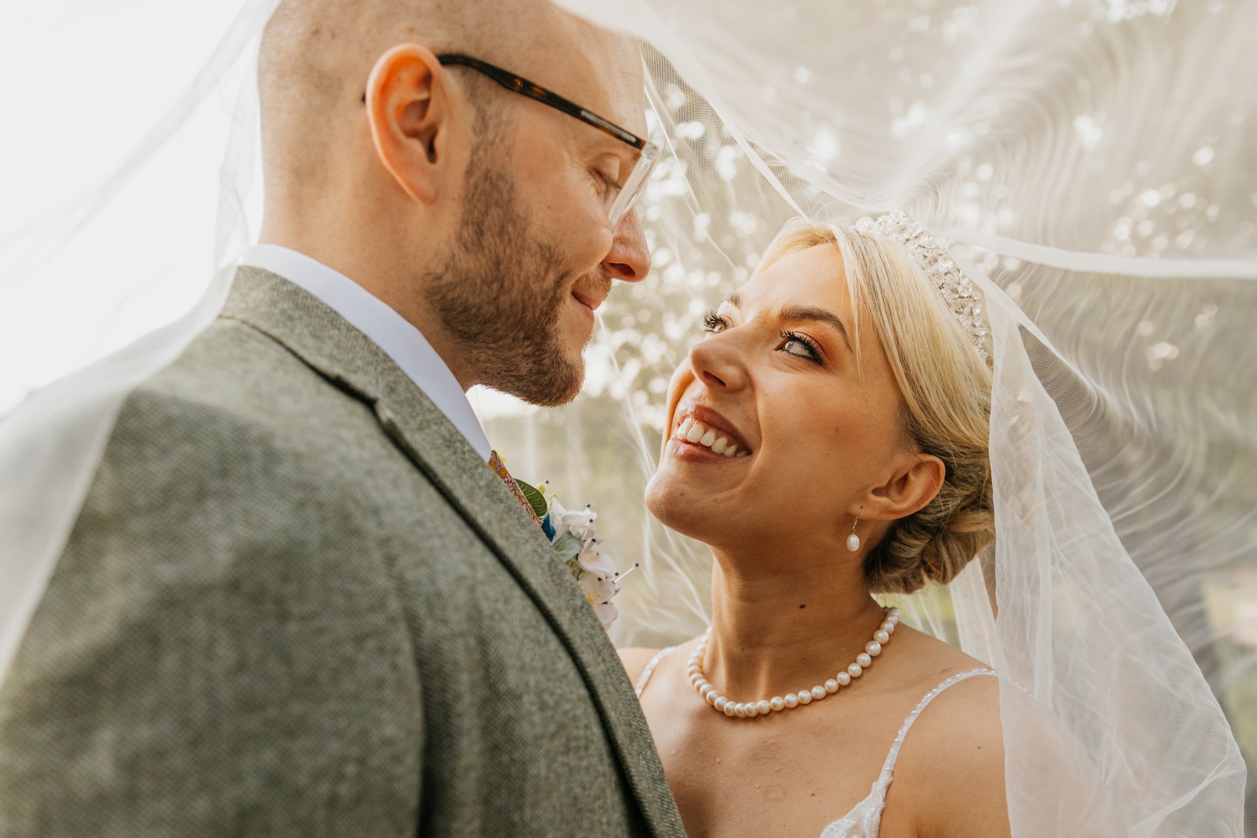A close-up of a bride and groom smiling and looking into each other's eyes under a wedding veil outdoors.