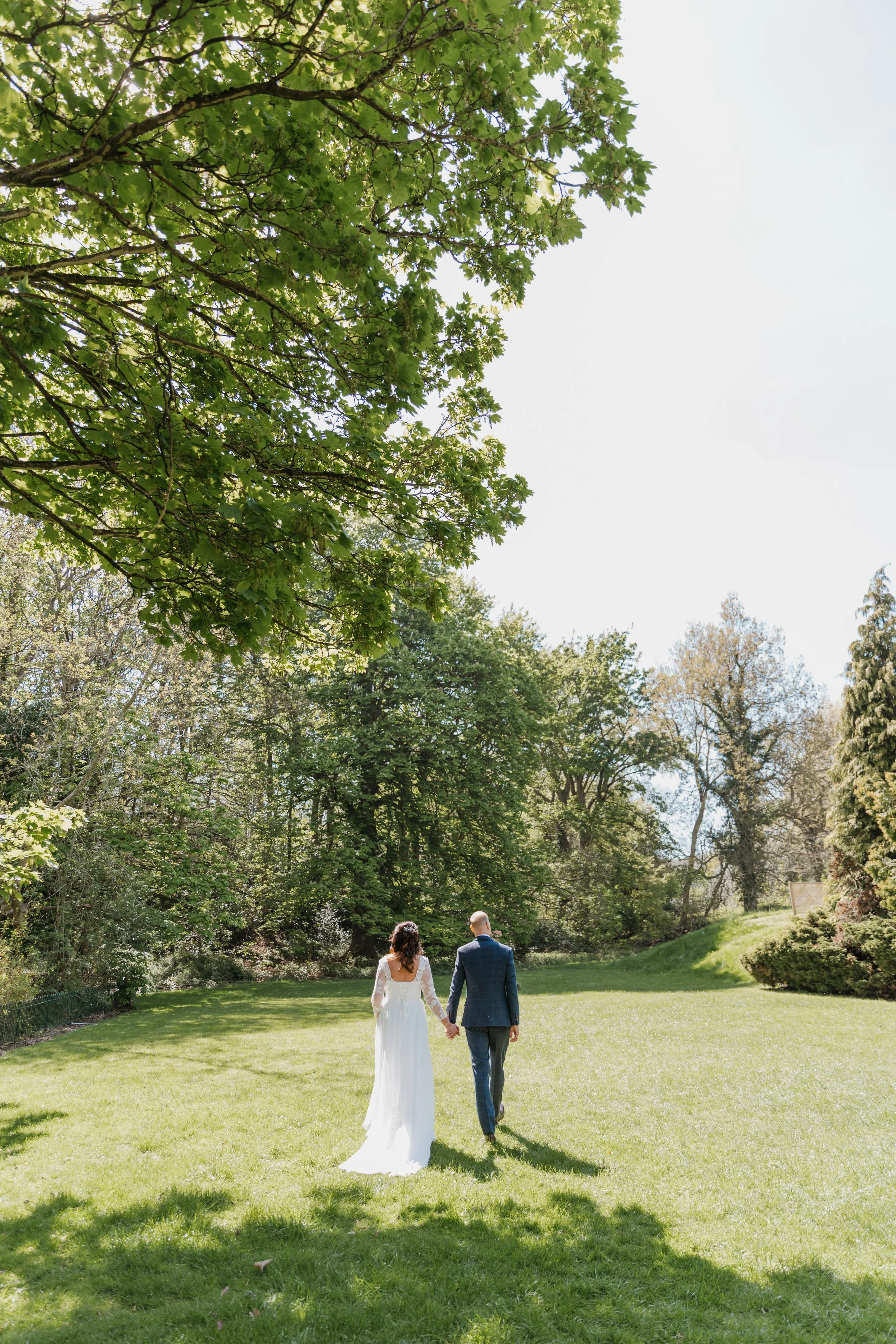 A bride and groom walking hand in hand on a grassy lawn, surrounded by tall trees under a bright sky.