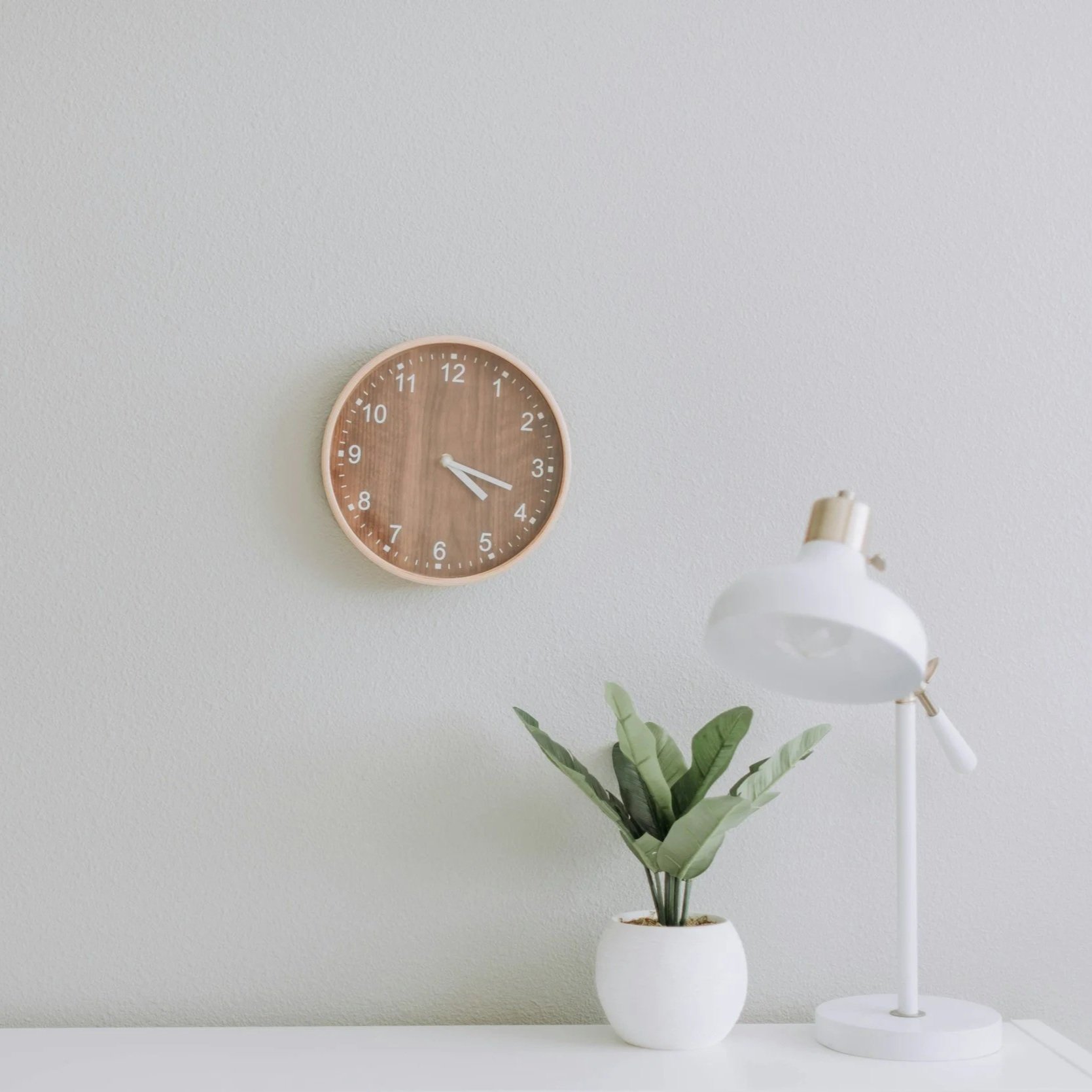 A wall clock showing 3:19 next to a white lamp and a potted green plant on a white surface against a light-colored wall.