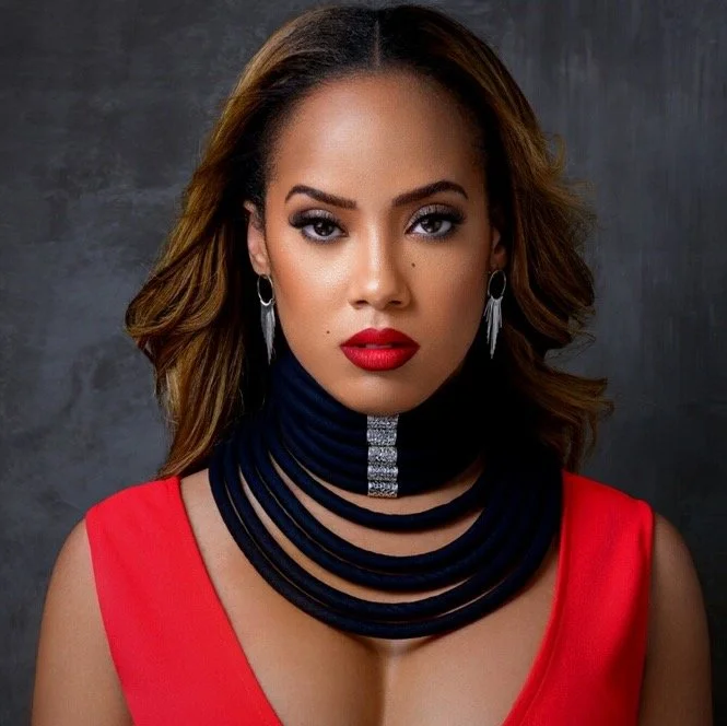 A woman with wavy brown hair, bold makeup, and red lipstick wearing a red sleeveless top and layered black necklace with a decorative silver stripe, standing against a dark textured background.