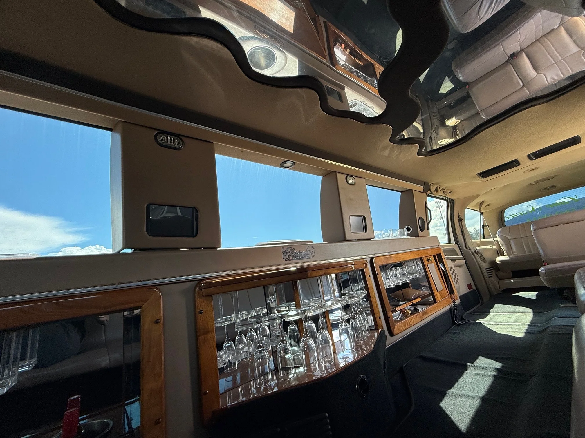 Interior of a vehicle with a large mirror on the ceiling, beige seats, and a glassware cabinet filled with glasses, with a view of a blue sky outside the window.
