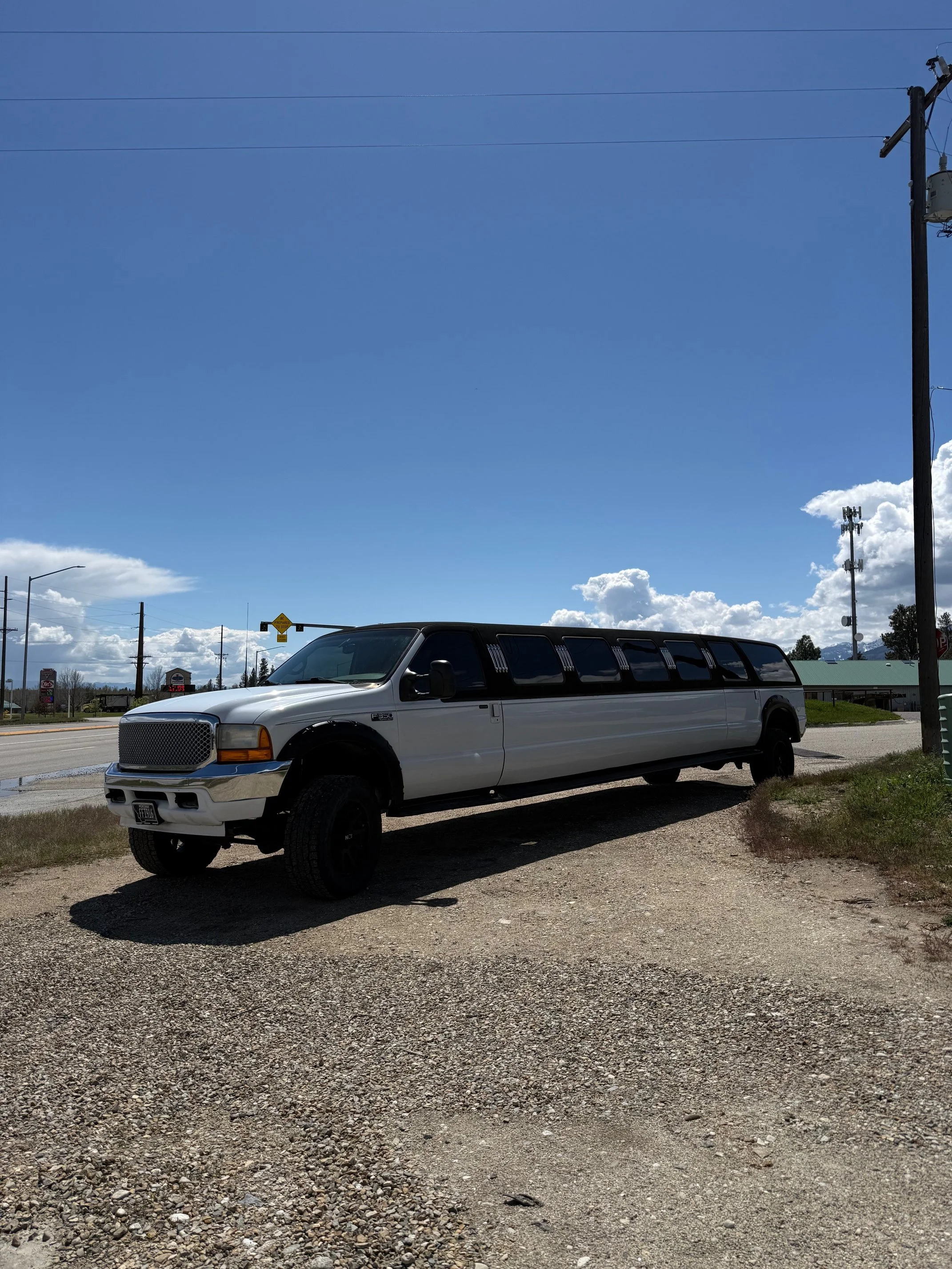 A white limousine parked on a gravel lot under a partly cloudy sky with utility poles and a street sign in the background.