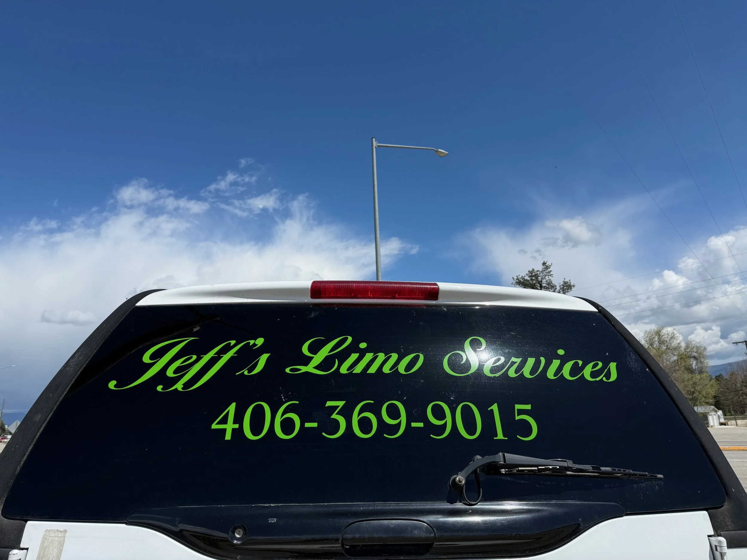 The back of a vehicle with a sticker that reads 'Jeff's Limo Services' along with a phone number. The vehicle is parked outdoors under a partly cloudy sky with a streetlamp visible.