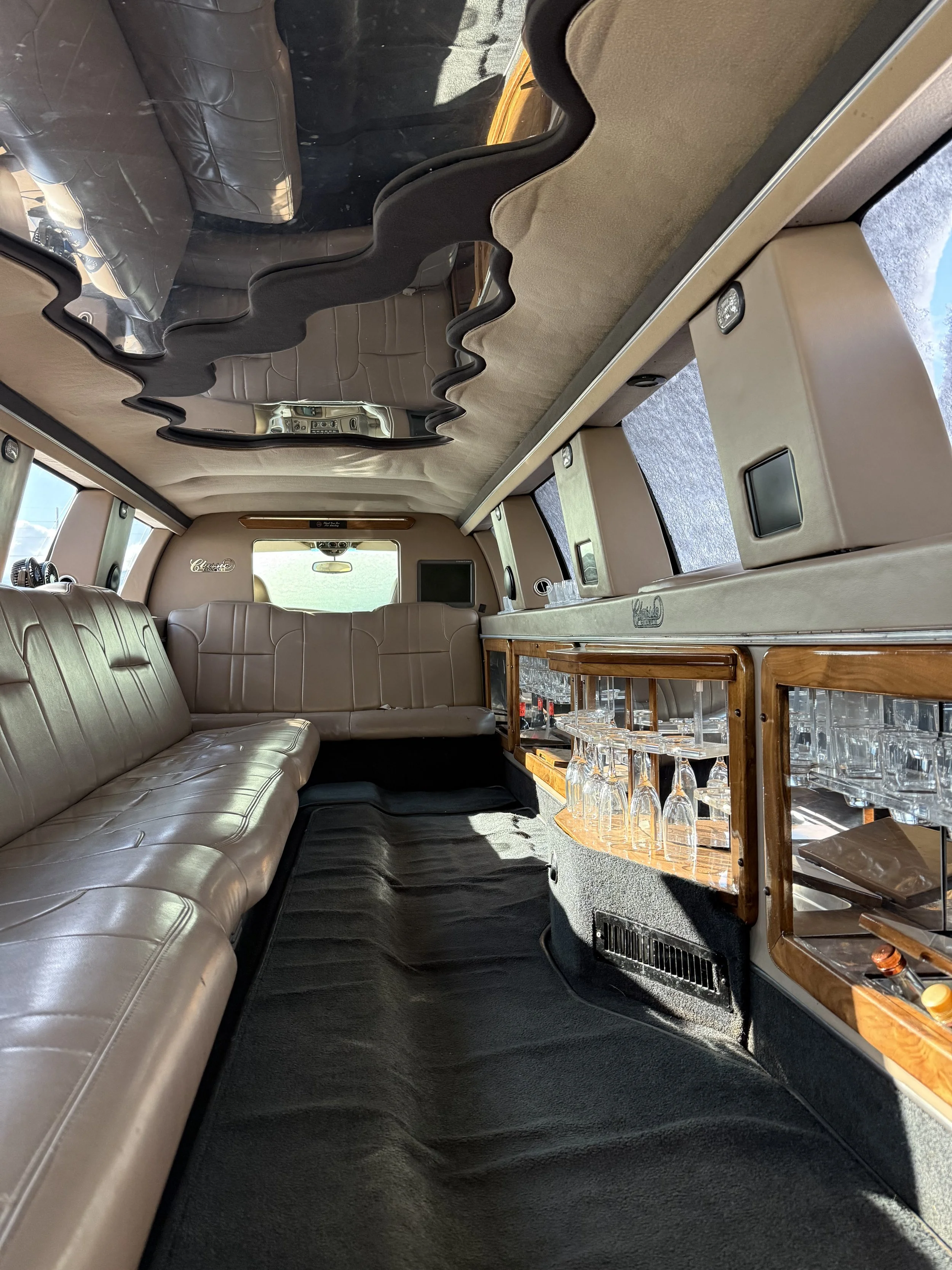 The interior of a vintage bus or camper van with a beige leather bench seat on the left and storage with glassware on the right. There is a mirror on the ceiling reflecting part of the room and a window at the back showing an outdoor scene.