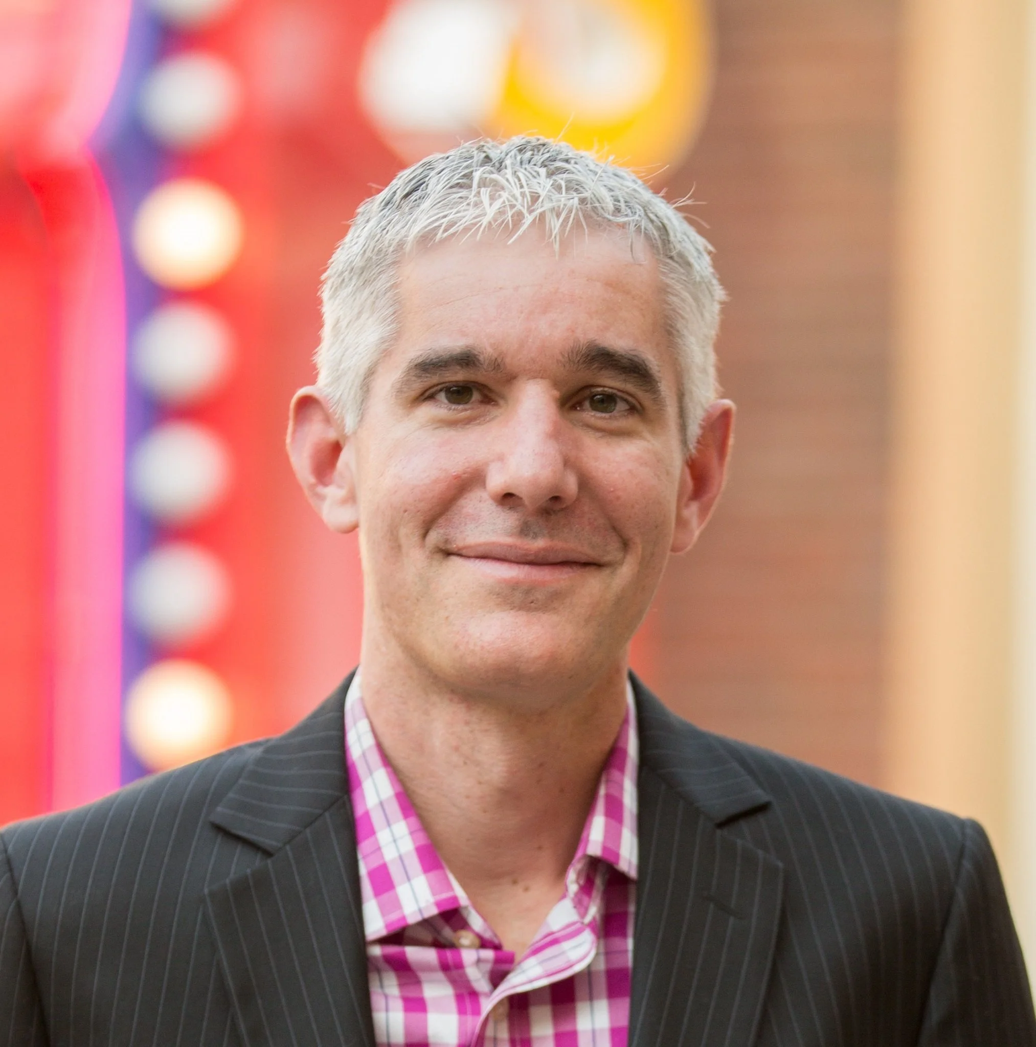Close-up of a man with short, gray hair wearing a dark pinstripe suit and a pink plaid shirt, smiling at an event with colorful blurred background lights.