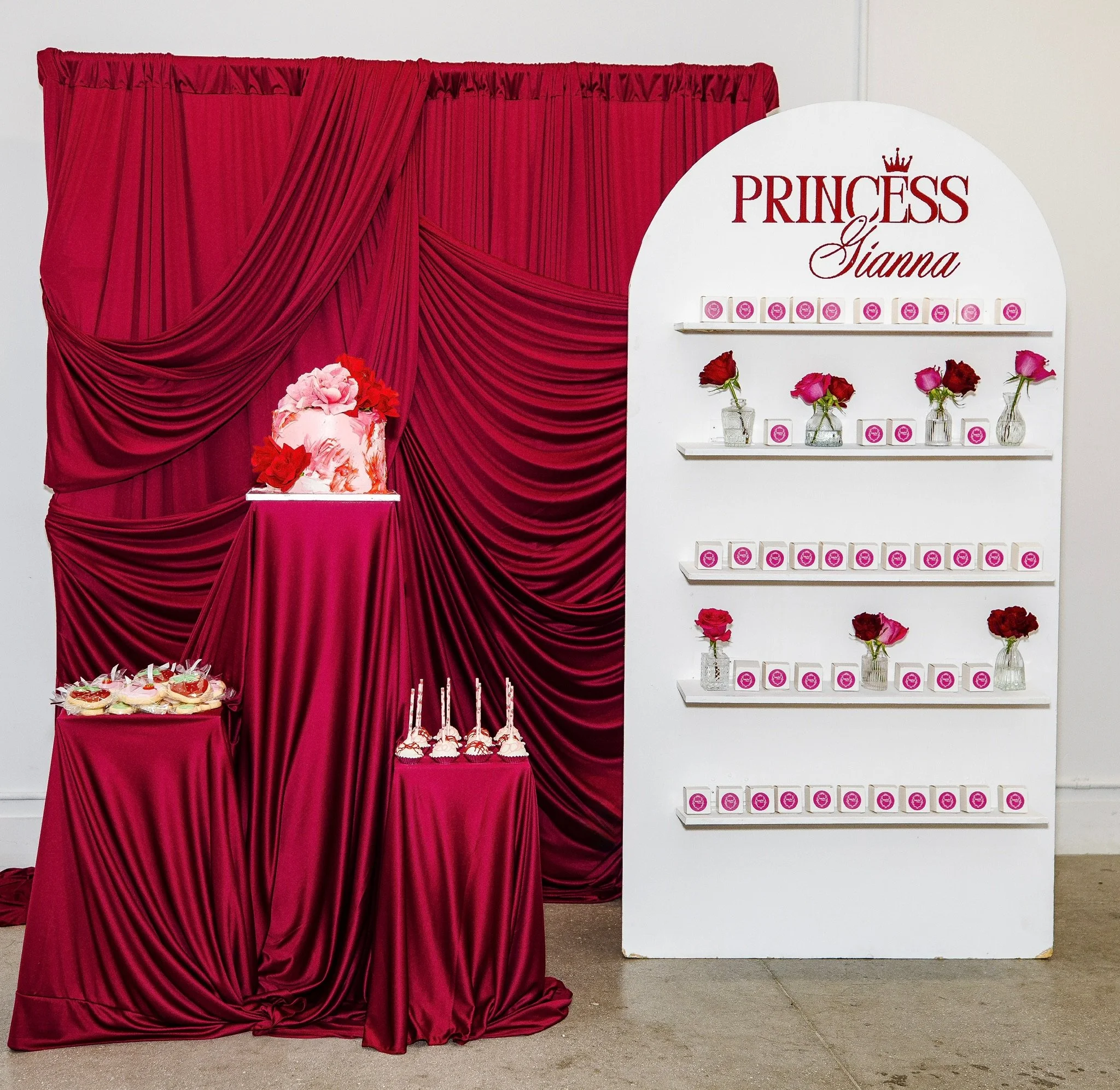 Decorative display with a red curtain backdrop, pink and red flowers, and a tutu on a small table, featuring a white display board with the text 'Princess Gianna' and pink-themed items, likely at a birthday party.