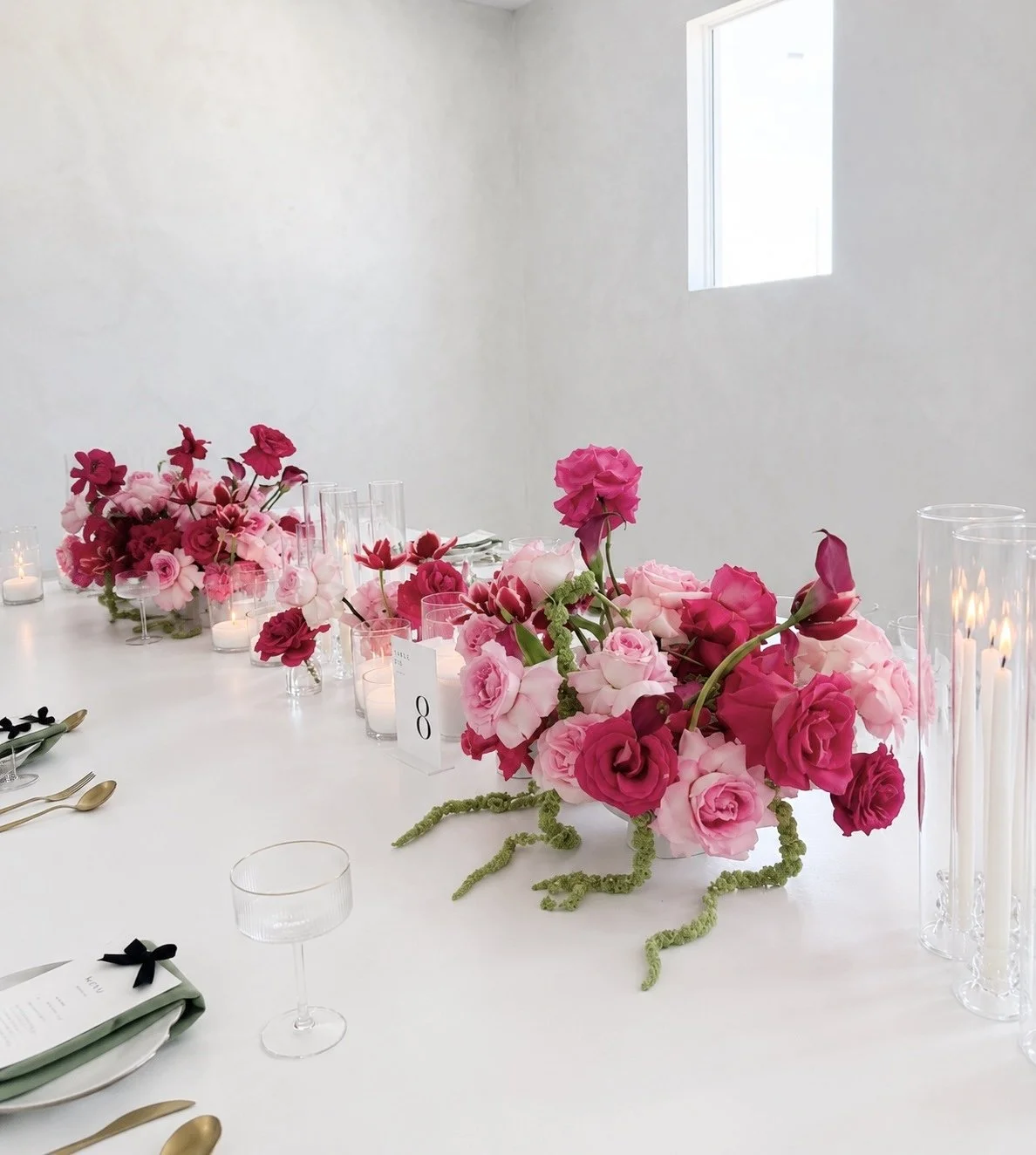 Elegant table setting with pink and red floral arrangements, candles, and gold and black utensils in a bright, minimalistic room with a small window.