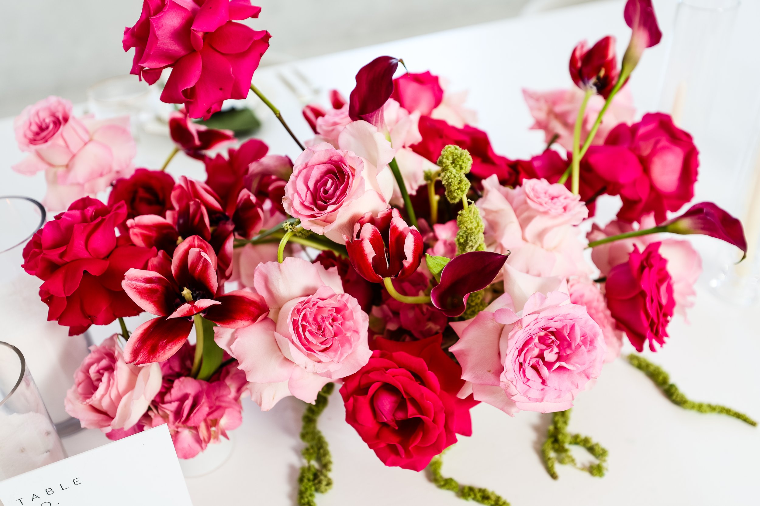 A bouquet of various pink and red flowers, including roses and calla lilies, with green stems and accents, arranged on a white surface.