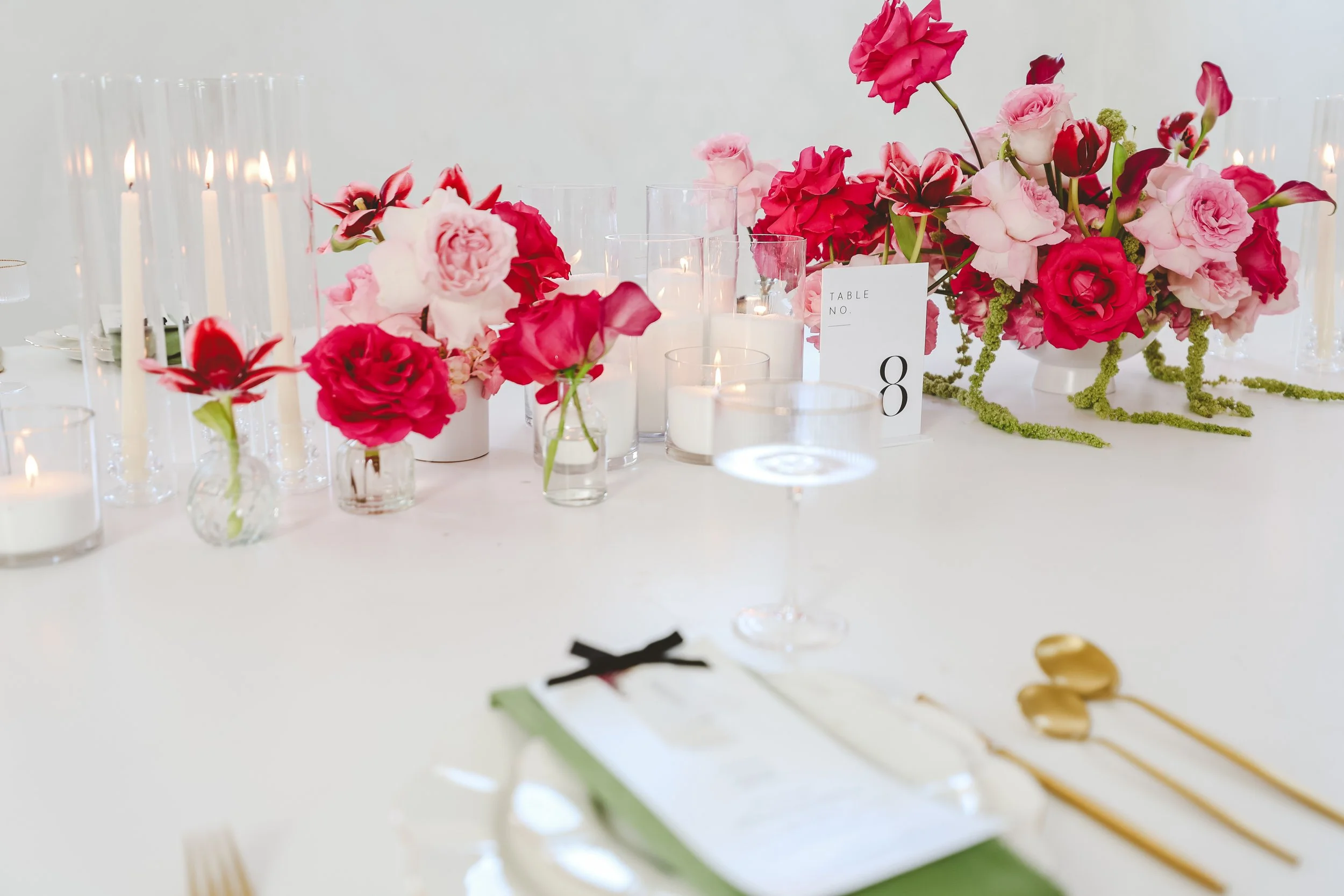 A table decorated with pink and red flowers, candles, and a white card with 'TABLE NO. 8' written on it, likely at a wedding or special event.