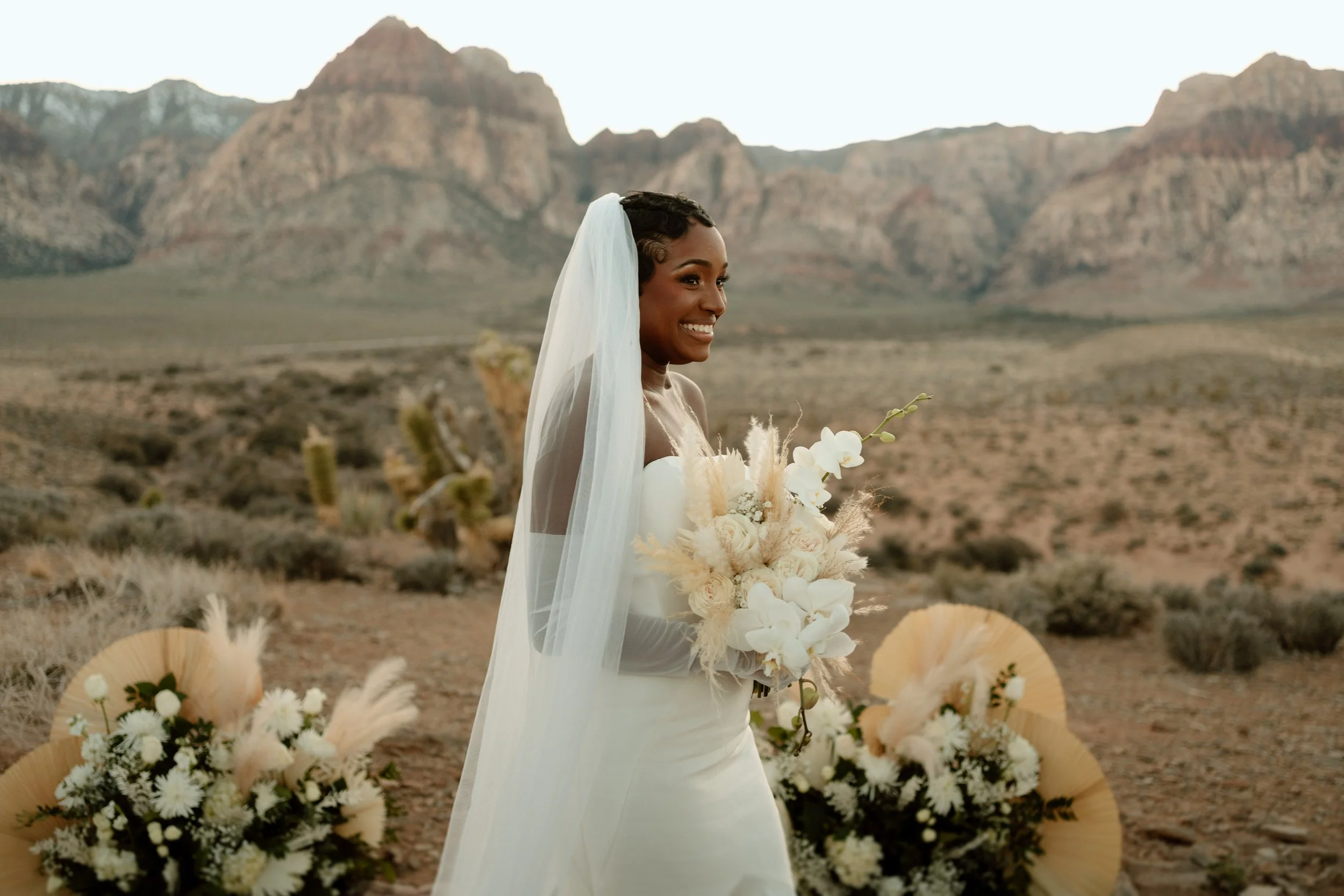 A smiling woman in a white wedding dress and veil holding a bouquet of white flowers in a desert landscape with mountains in the background.