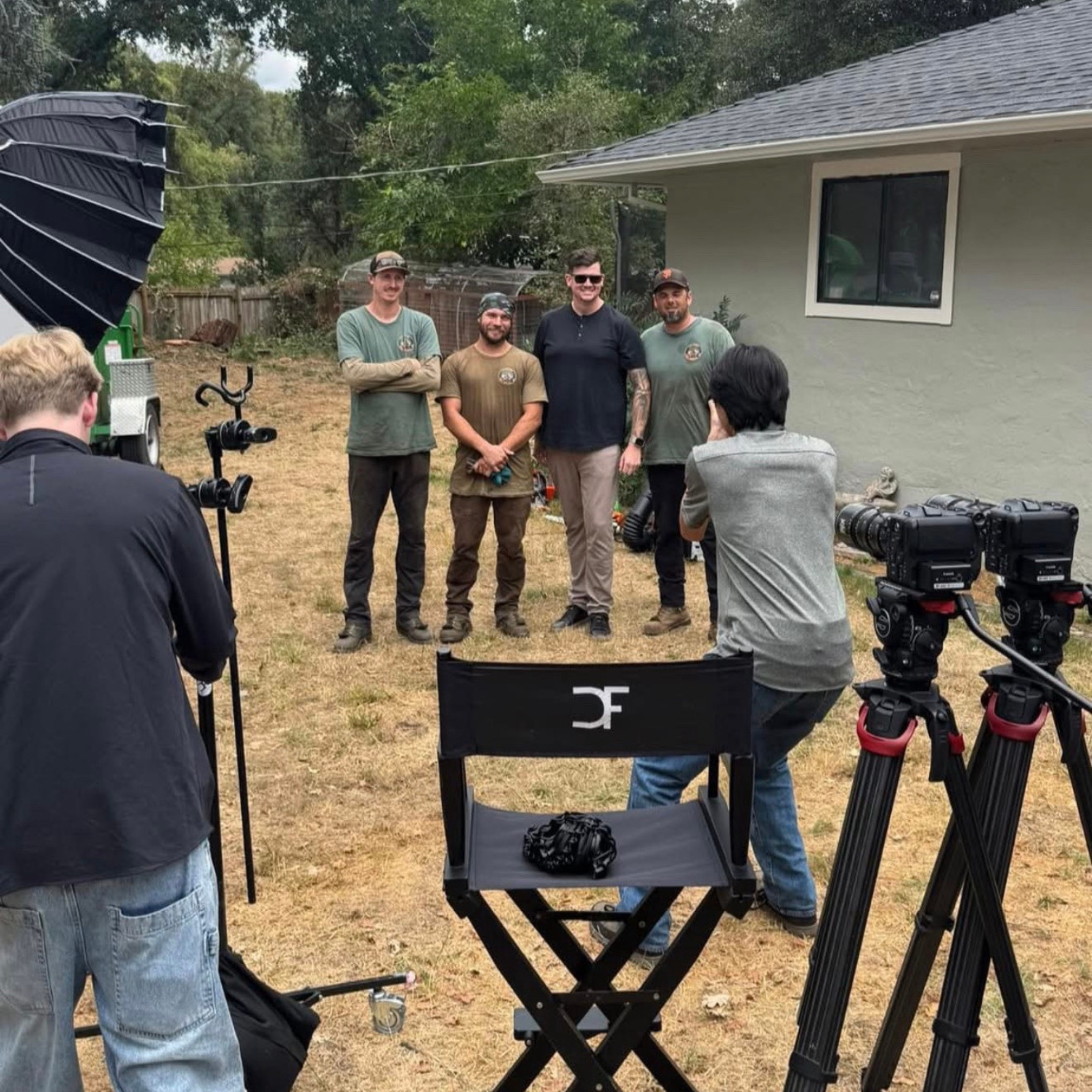 Four men stand side by side outdoors near a house, smiling for the photo during daytime. A photography or filming setup with cameras and lights is visible in the foreground.