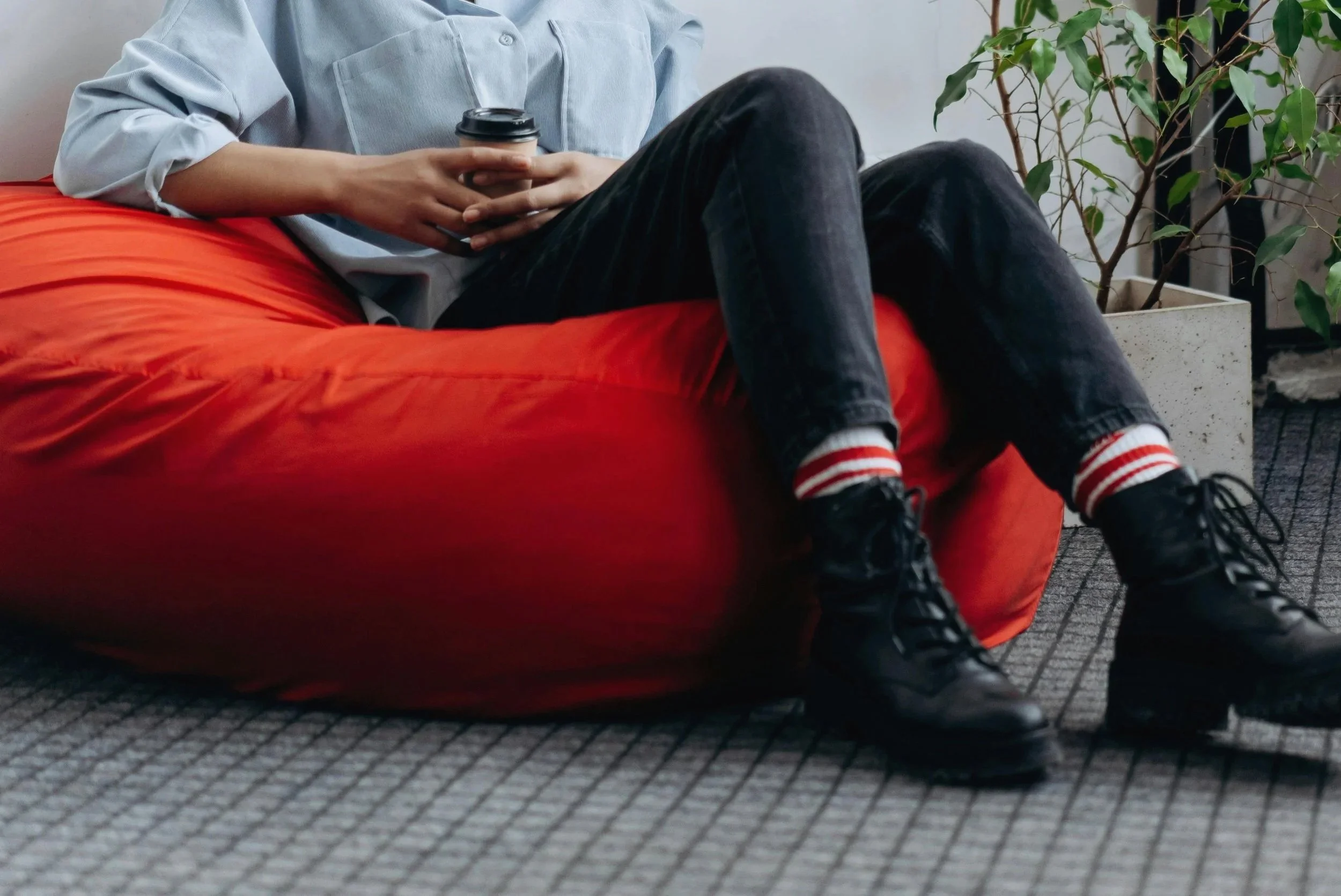 Person sitting on a red couch, holding a cup of coffee, wearing a light blue shirt, black pants, and striped socks, near a potted plant in an indoor setting.