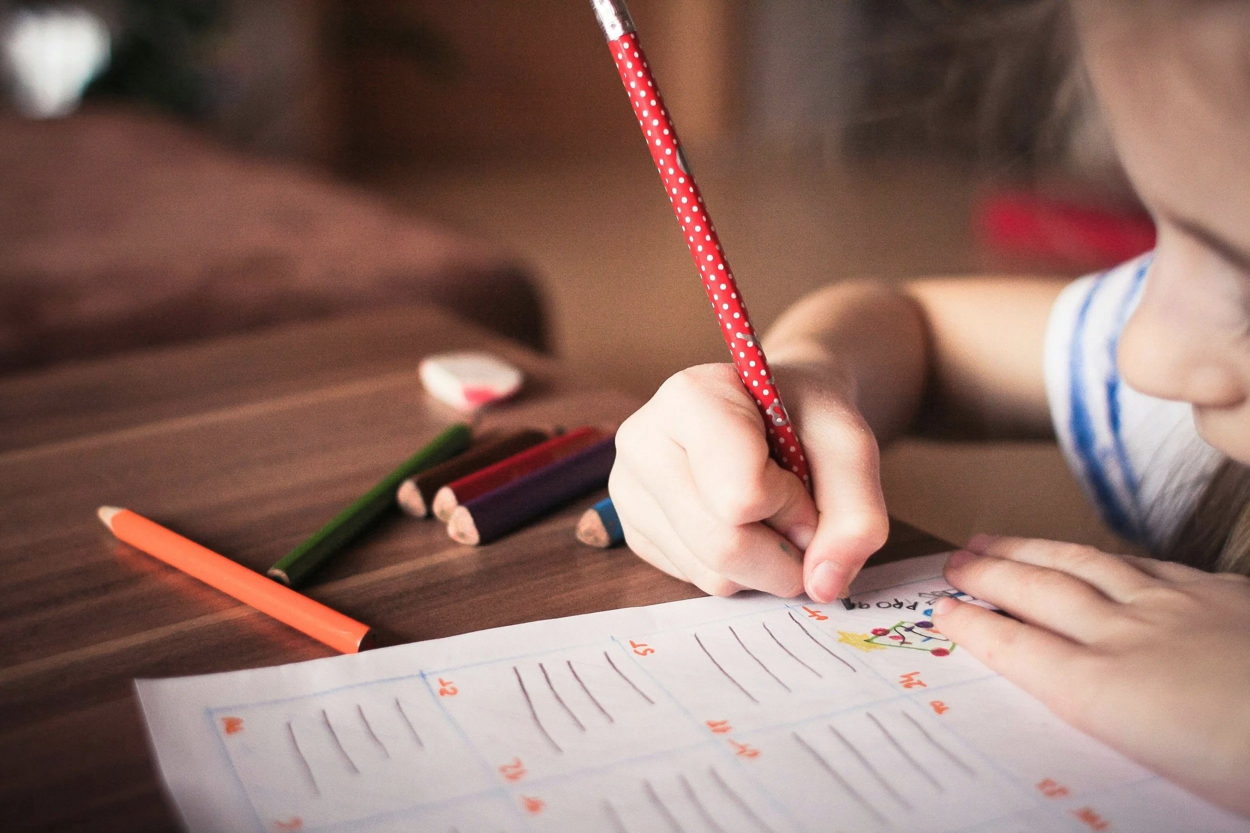 A child's hand holding a red polka dot colored pencil, writing on a lined notebook, surrounded by colorful pencils on a wooden surface.