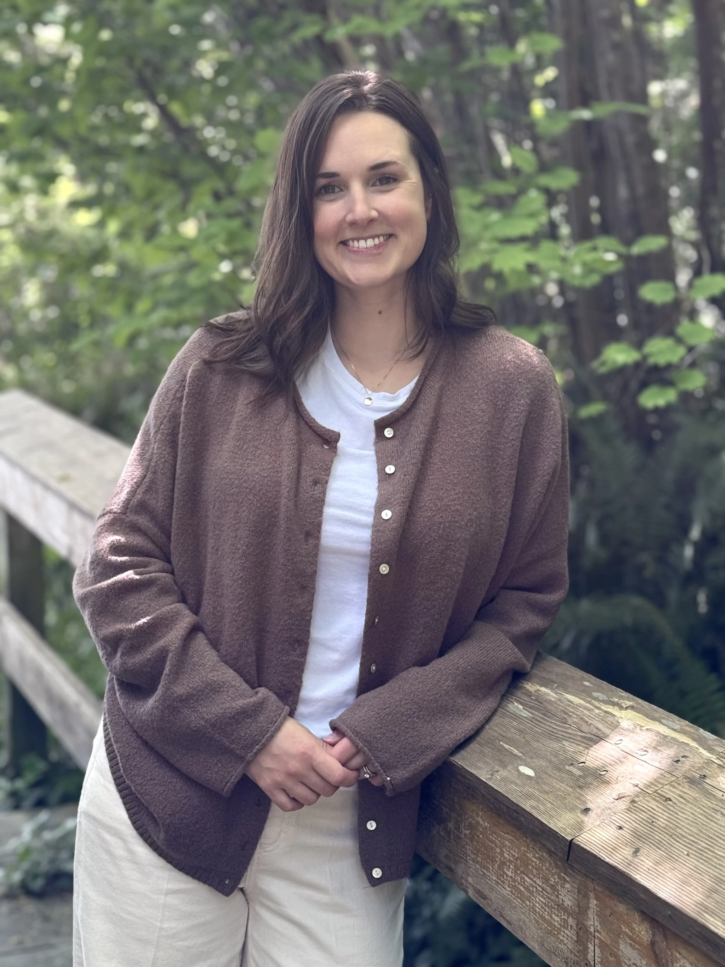 A woman standing outdoors on a wooden bridge, smiling, with green trees in the background, wearing a brown cardigan over a white shirt.