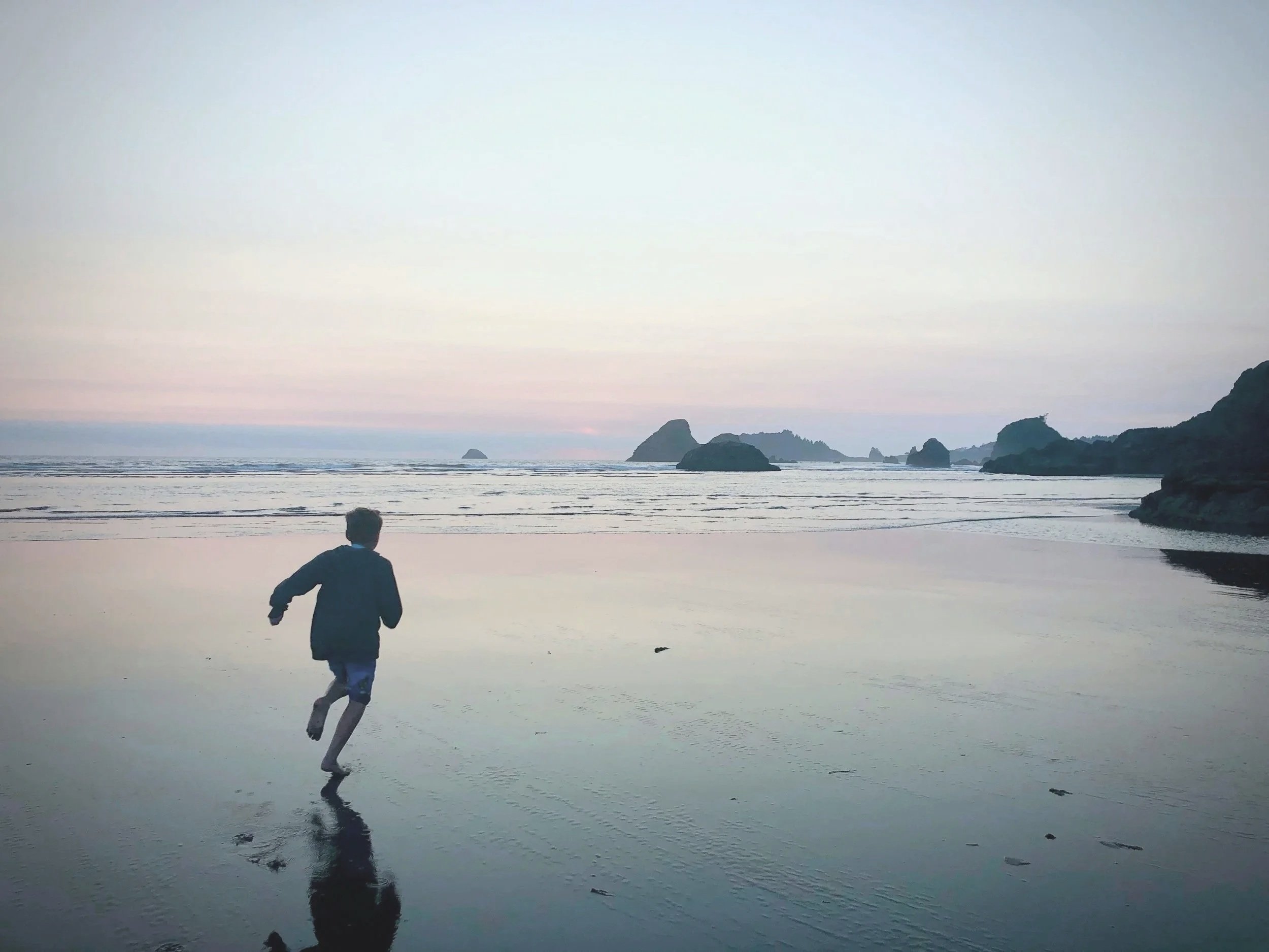 A person running on a beach during sunset, with rocks and small islands in the background, and the sky transitioning from light blue to pink.