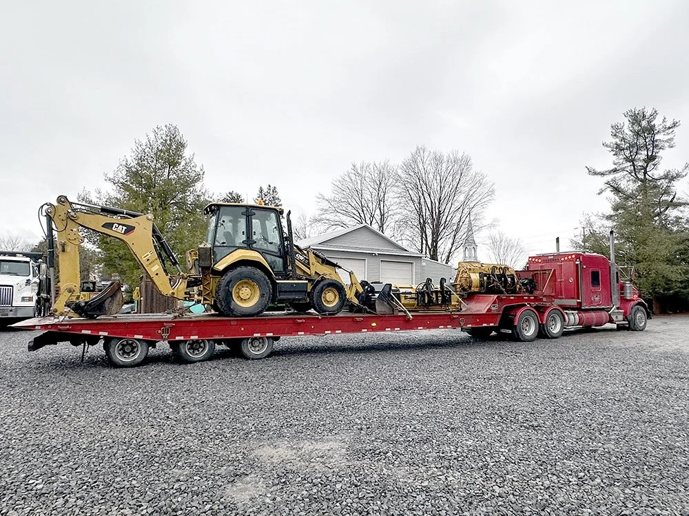 A large red flatbed truck transporting construction equipment, including a yellow backhoe loader and excavator attachments, parked on gravel with trees and buildings in the background.