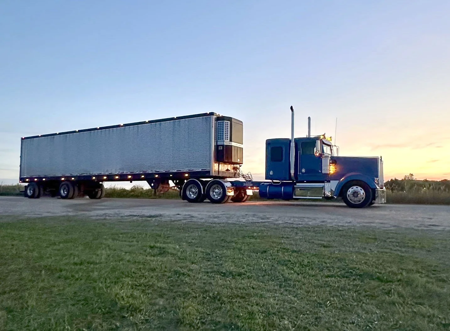 A blue semi-truck with an empty trailer parked on a grassy area during sunset.