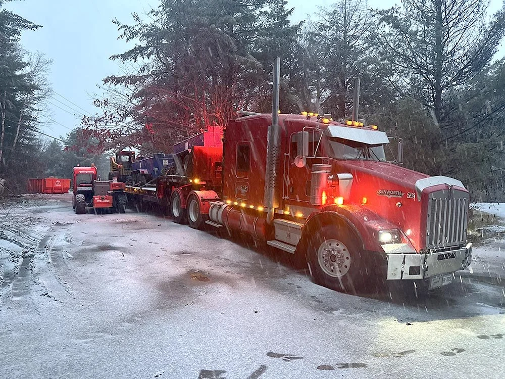 Large red semi-truck stuck in a snowy, icy road during rain, with snow plows and construction equipment nearby, surrounded by trees.