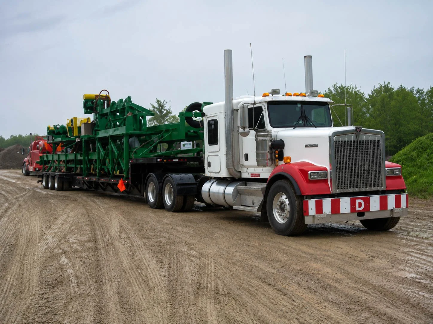 A semi-truck with a white cab and red accents is parked on a dirt road, hauling green machinery.