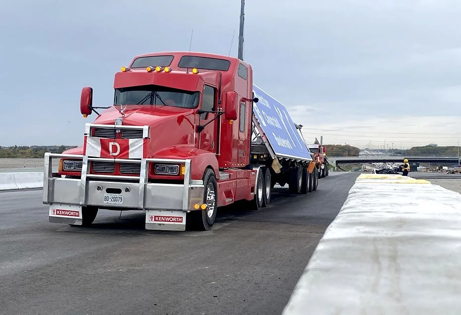 A red Kenworth semi-truck on a bridge or overpass, with a blue sign on its trailer, and construction workers visible in the background.