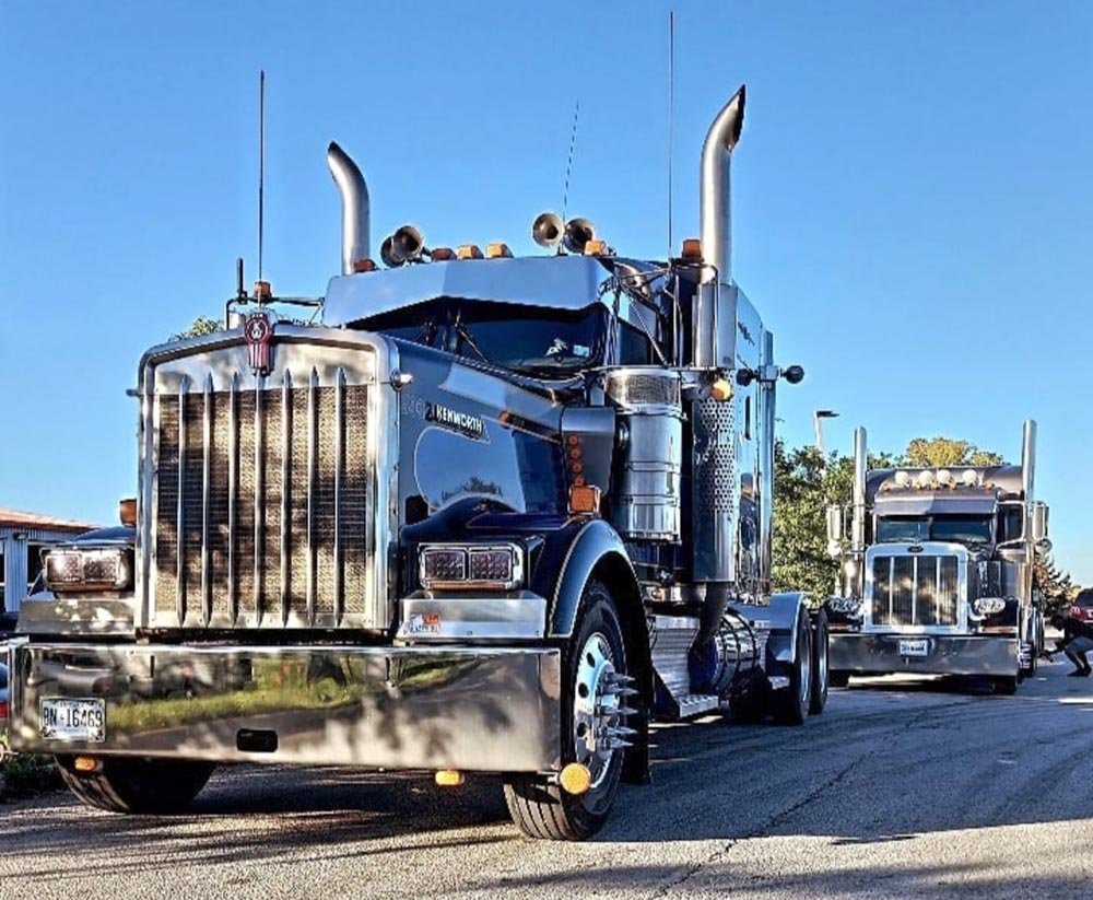 Two large black semi-trucks parked on a street during daytime with clear blue sky.