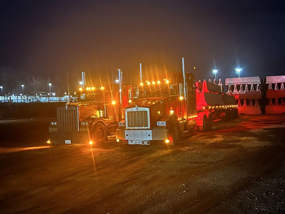 Two large semi-trucks parked side by side at night, illuminated by their orange warning lights, with construction barriers visible in the background.