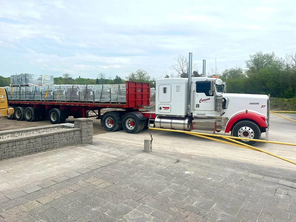 A large white and red semi-truck parked on a paved area, hauling a flatbed trailer loaded with wrapped pallets.