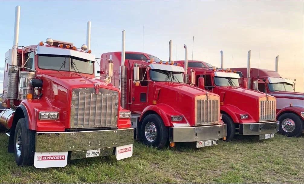 Line-up of red semi-trucks on grass field at sunset.